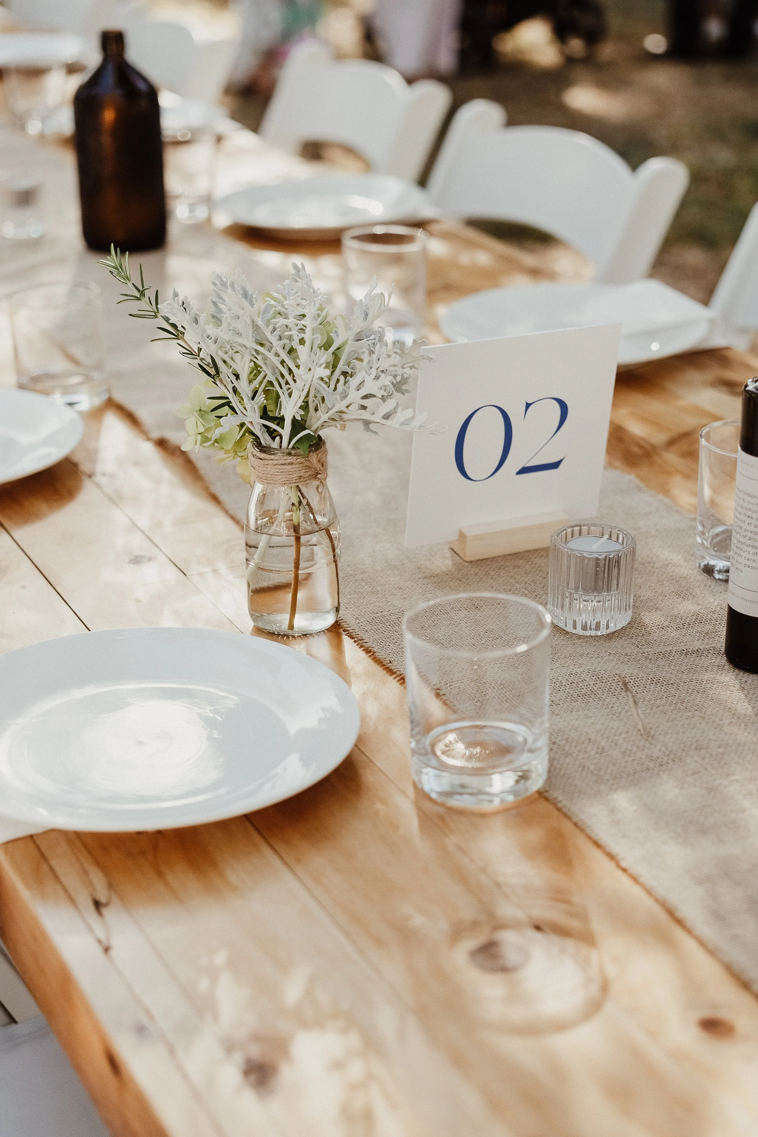 A table decorated for an event, featuring a white flower arrangement, table number 02, clear glasses, and white plates. The table appears to be made of wood with a beige table runner.