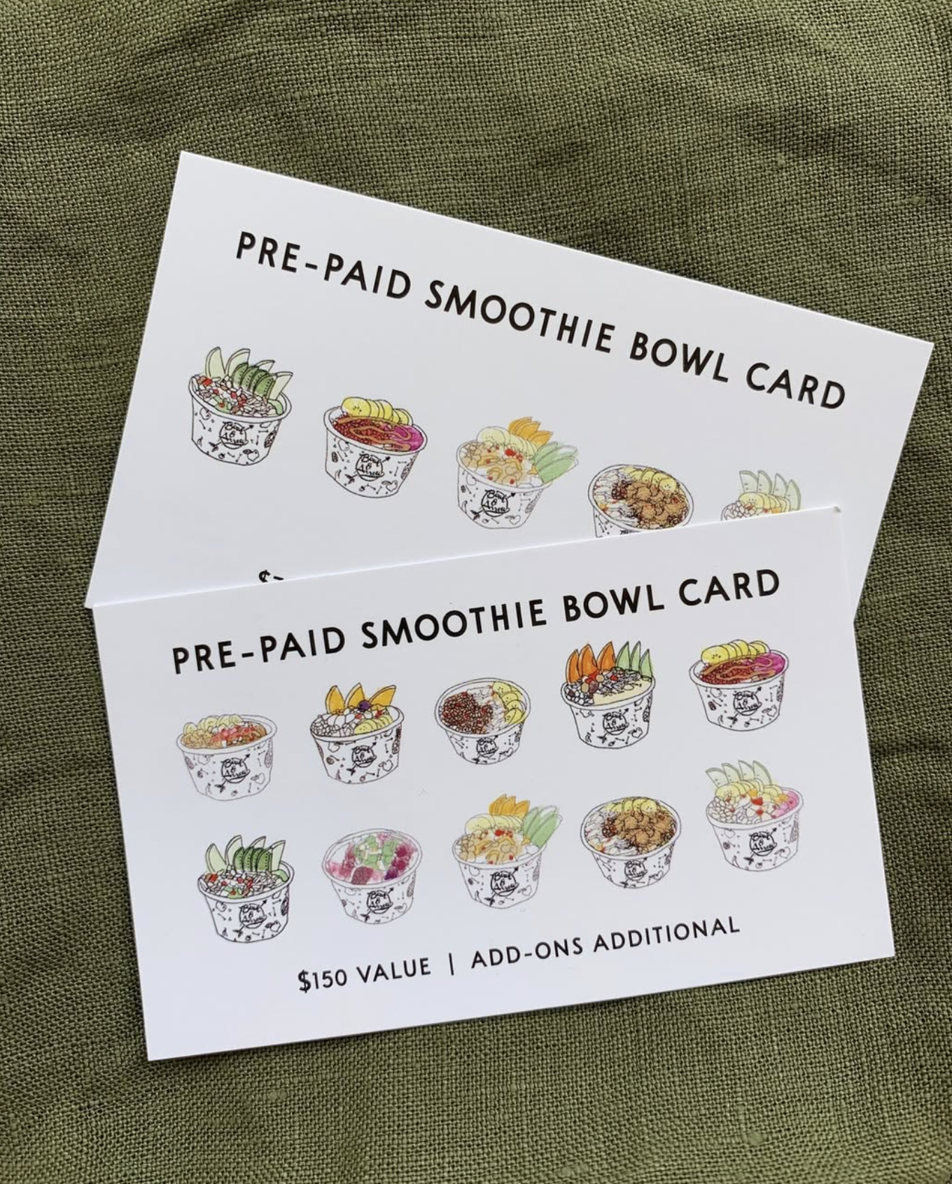 Two pre-paid smoothie bowl cards showing colorful illustrations of various smoothie bowls with toppings, placed on a green fabric surface.