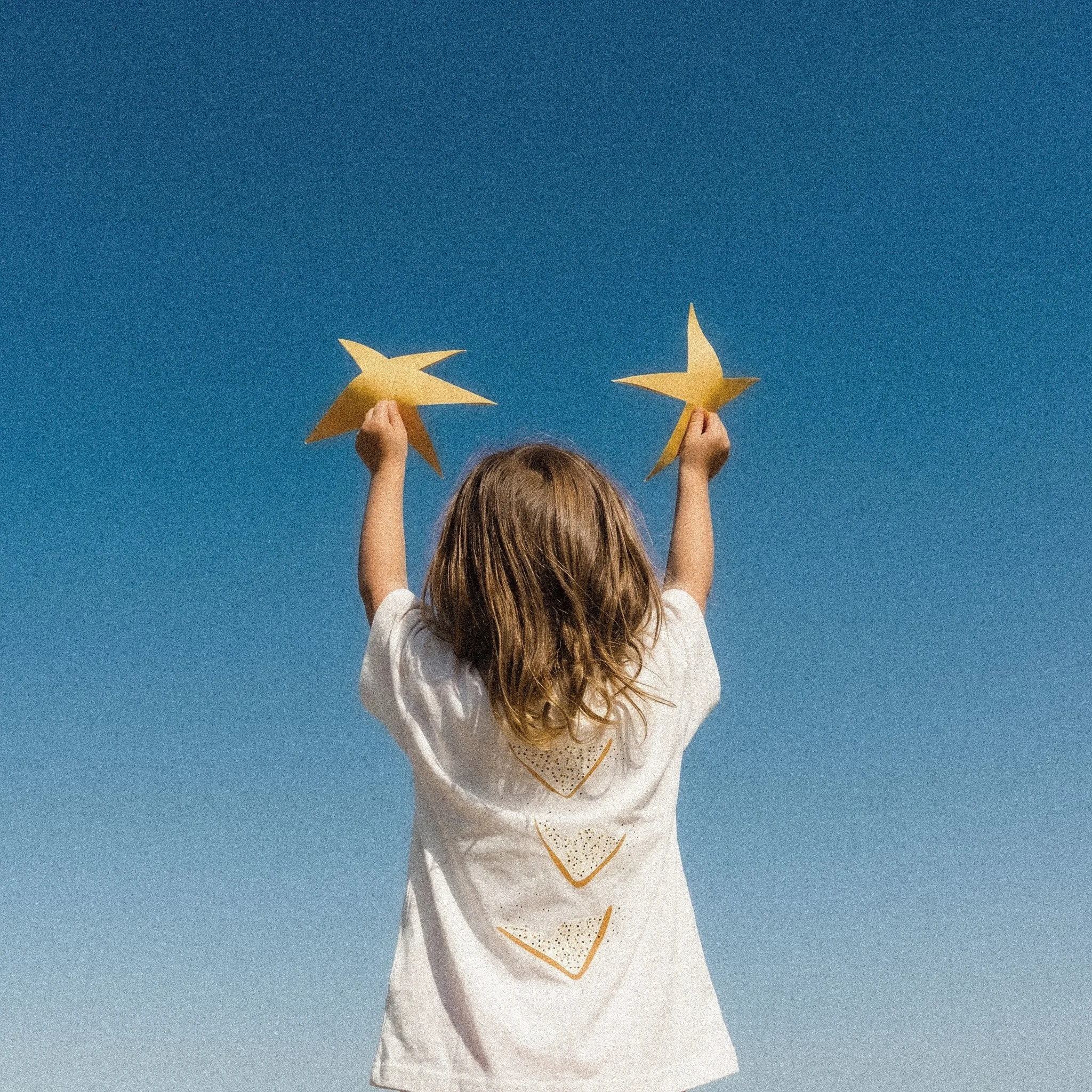 A child with long light brown hair holds gold paper stars against a clear blue sky.