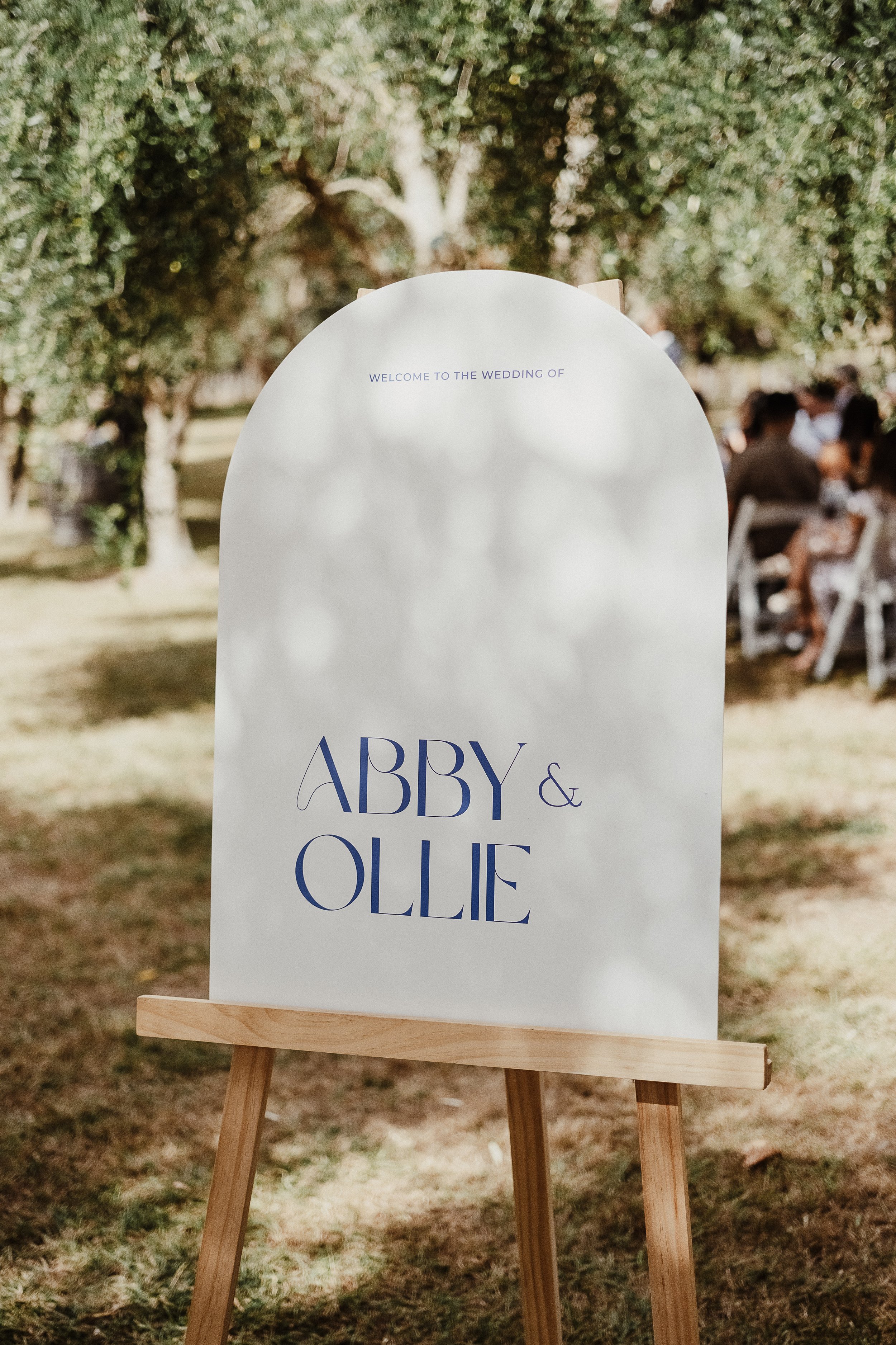 Wedding welcome sign with the names Abby and Ollie, displayed on an easel outdoors with trees and guests in the background.