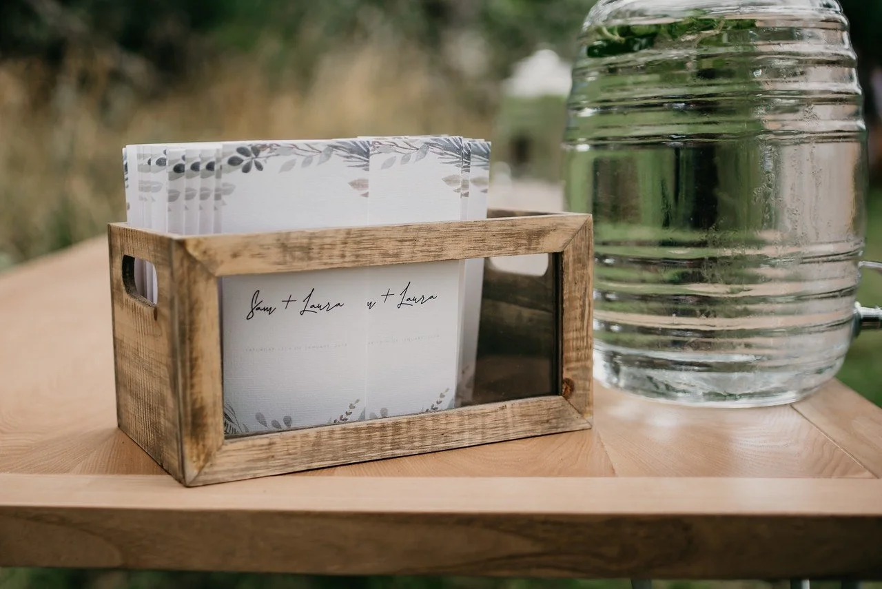 A wooden tray holds a glass jar of water and a small wooden frame containing stationery with a handwritten note saying 'Sam + Laura' and a decorated card with floral designs.