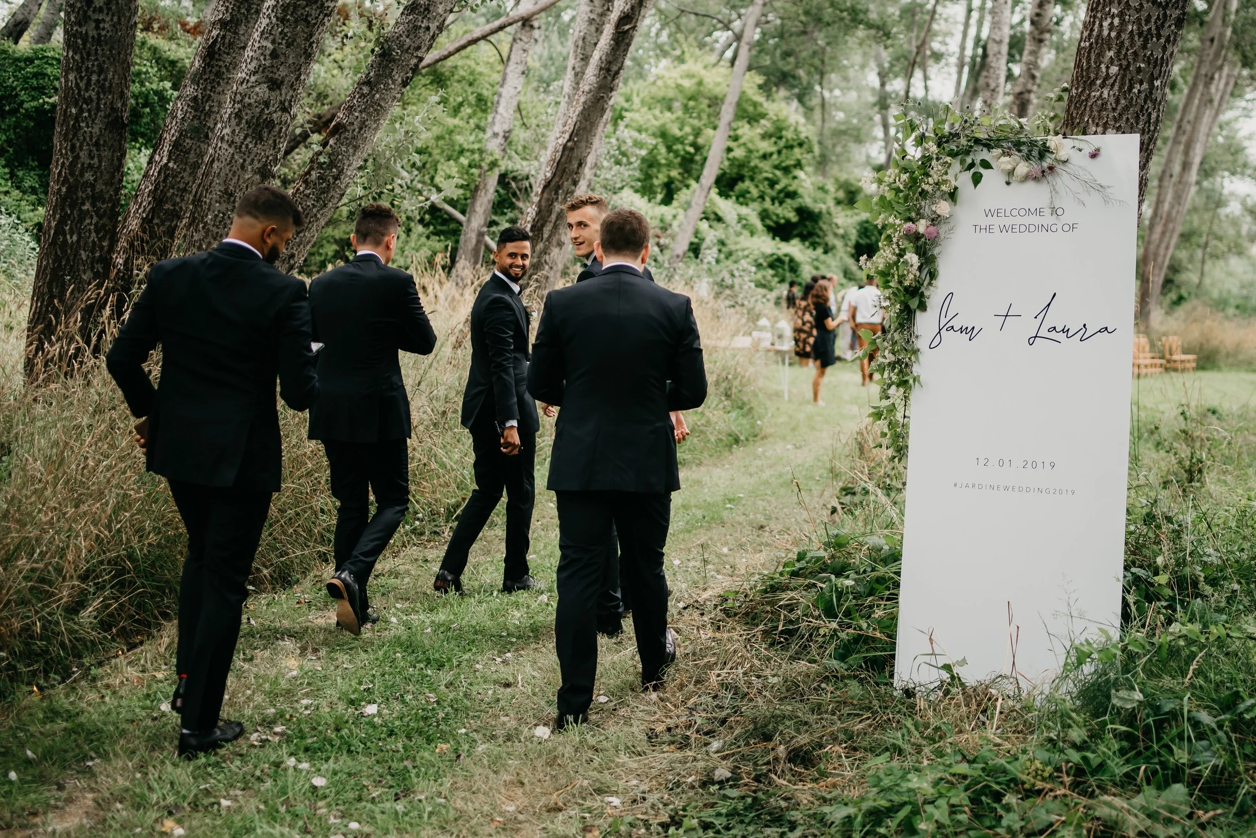 Group of men in black suits standing and talking near a wedding welcome sign outdoors in a wooded area.