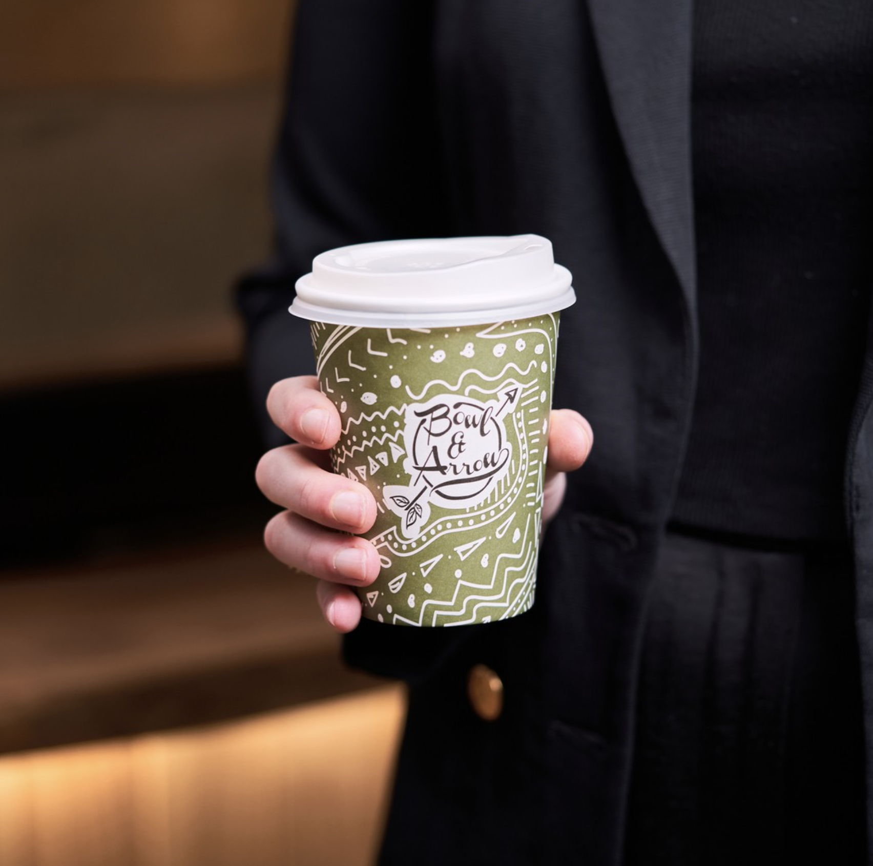 Person holding a takeout coffee cup labeled 'Bowl & Arrow' in a green and white patterned design, wearing a dark suit jacket.