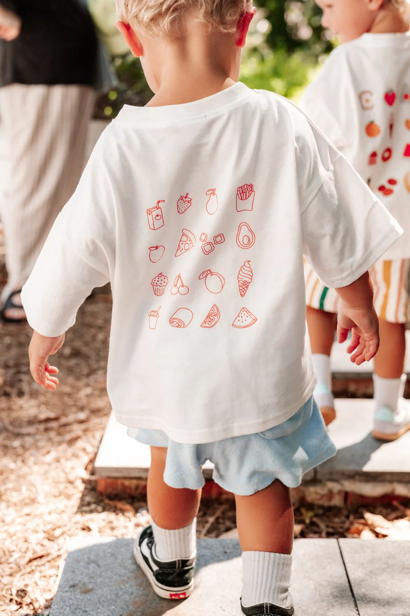 Child wearing a white T-shirt with red line drawings of various food items like pizza, strawberries, and fries, along with other snacks and drinks, standing outdoors.