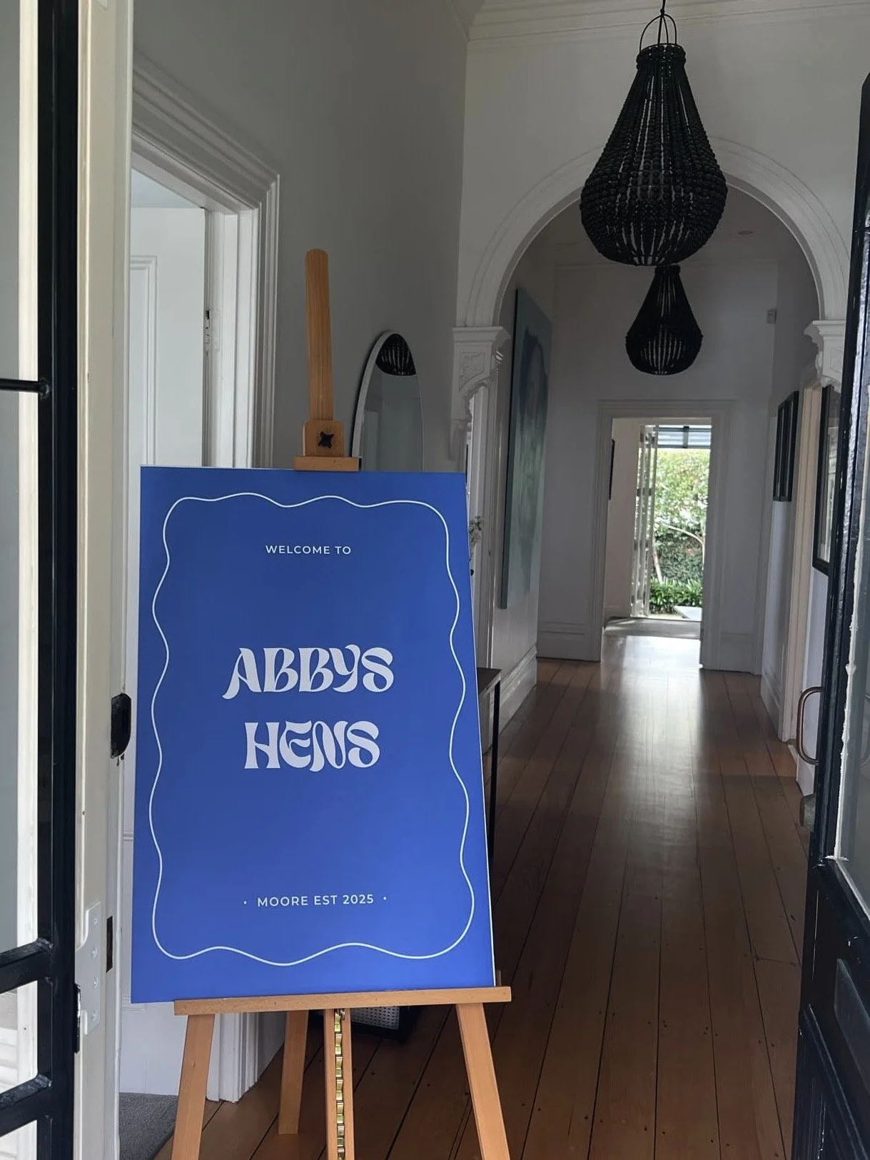 A blue sign on an easel inside a house reads 'Welcome to Abby’s Hems, Moore Est 2025.' The house has wooden floors, white walls, and a hallway with a doorway leading to a brightly lit outdoor area.