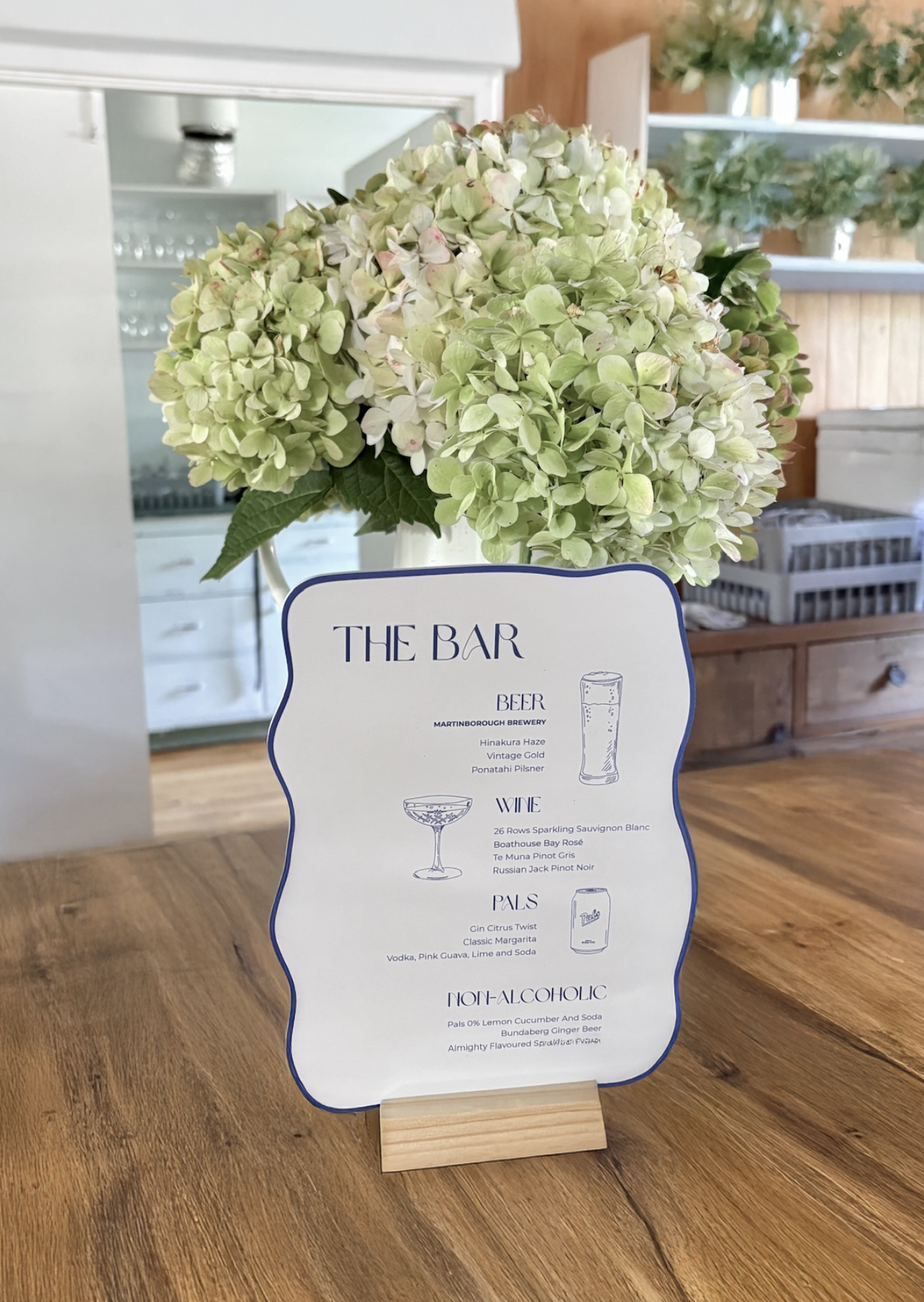 A menu card on a wooden table with a hydrangea flower arrangement behind it. The menu lists drinks such as beer, wine, pals, and non-alcoholic beverages.
