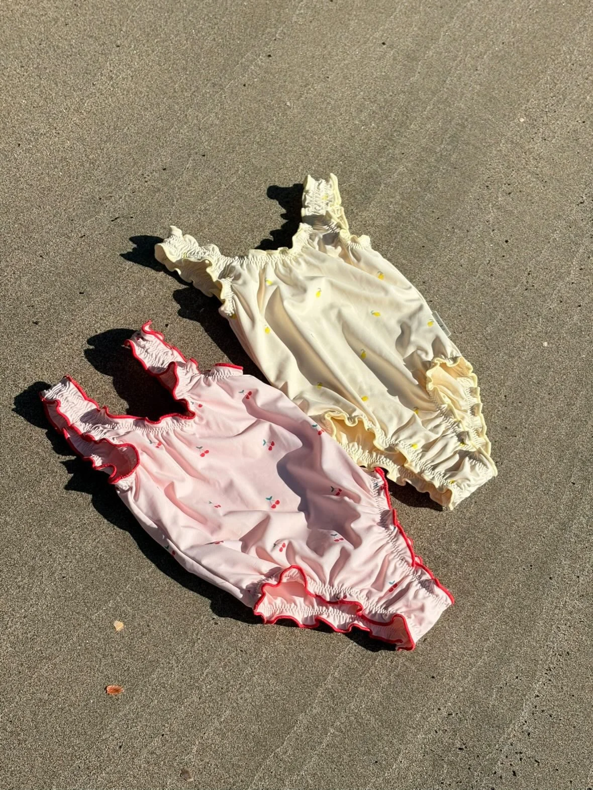 Two pairs of children's ruffled swimsuits, one pink with cherry patterns and red ruffles, the other cream yellow with small yellow dots and ruffles, lying on concrete ground.