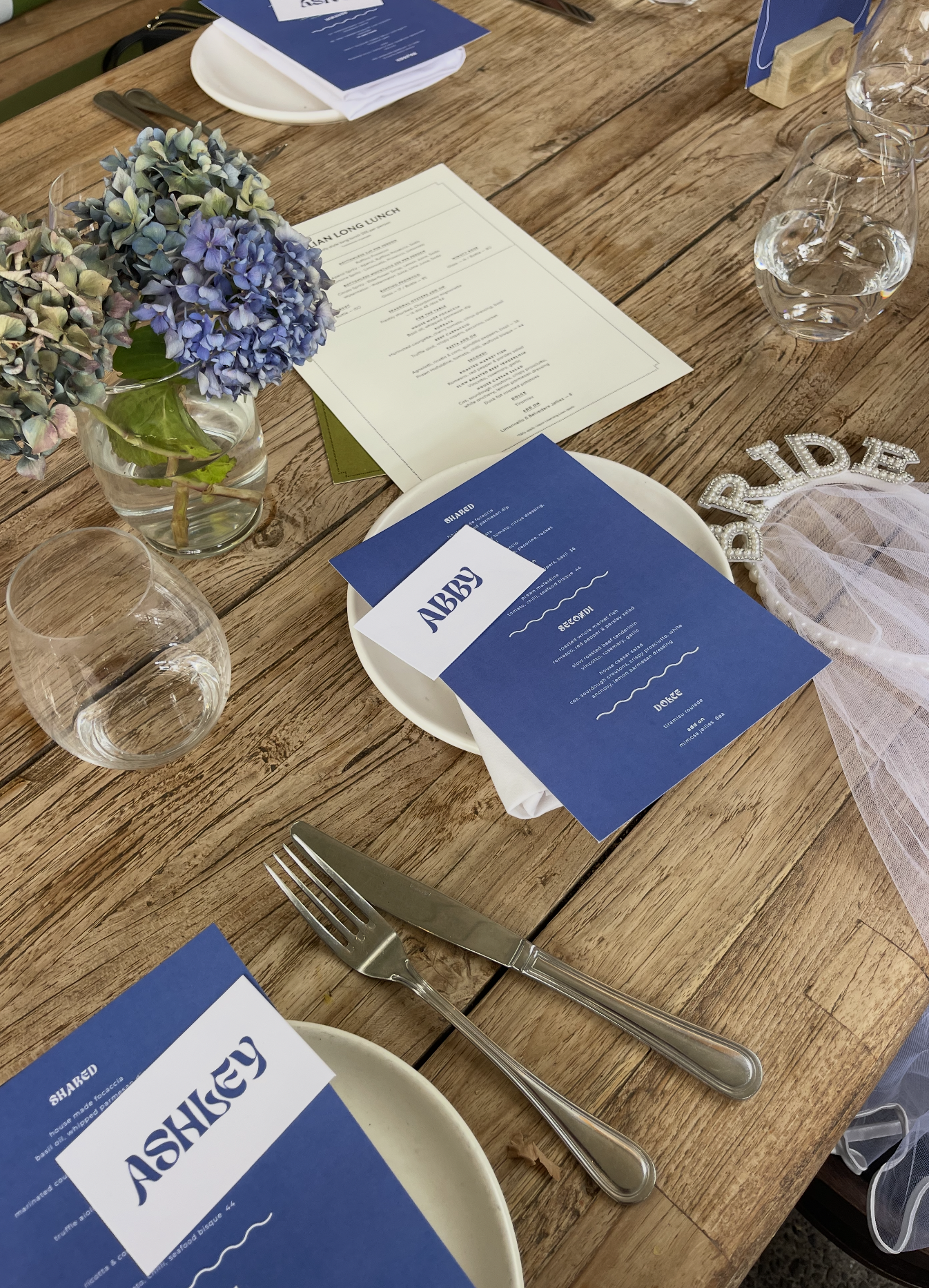 A wooden table set for a meal with a flower arrangement, labeled menus, glasses of water, silverware, and decorative items. The menus and items on the table suggest a formal dining event.