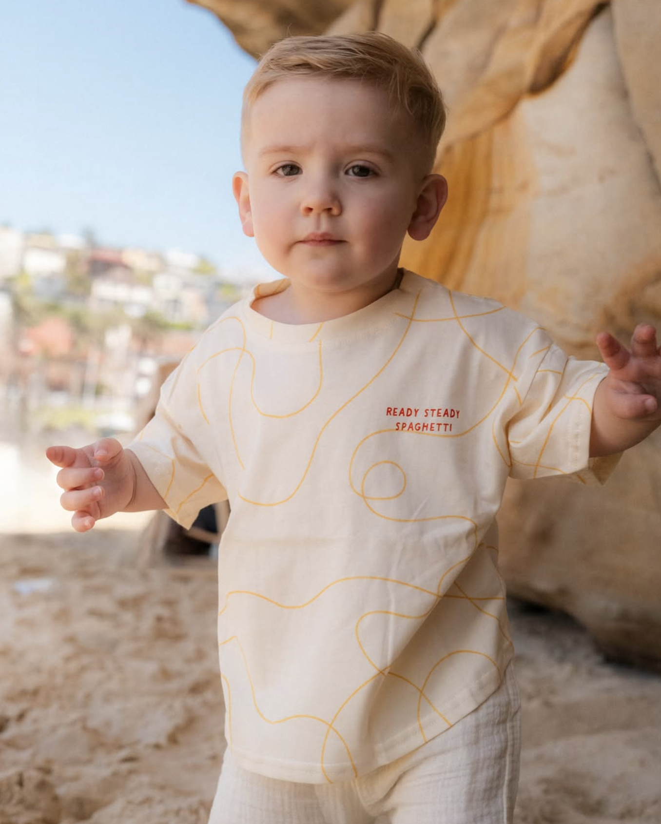 A young boy with blonde hair, wearing a cream-colored shirt with orange swirl patterns and the words "READY STEADY SPAGHETTI" on it, standing outdoors on a rocky surface near a large rock formation, with a blurred background of buildings and a clear 