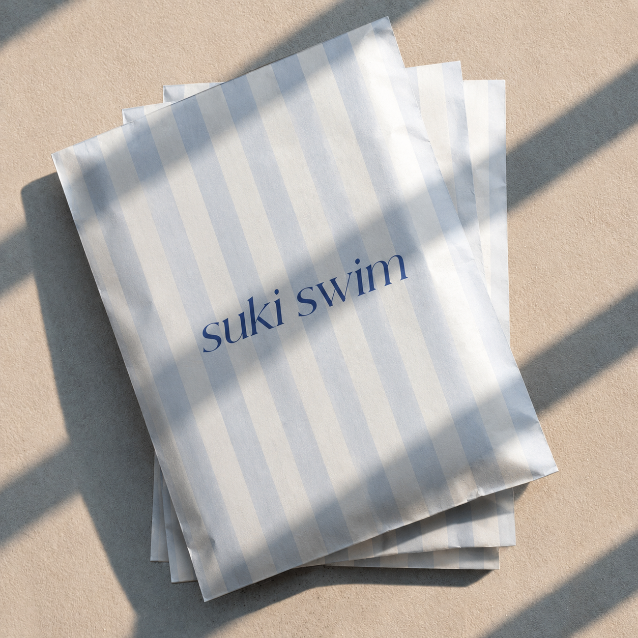 Paper bags with blue and white vertical stripes and the text 'suki swim' printed in blue, lying on a beige surface with shadows across the bags.