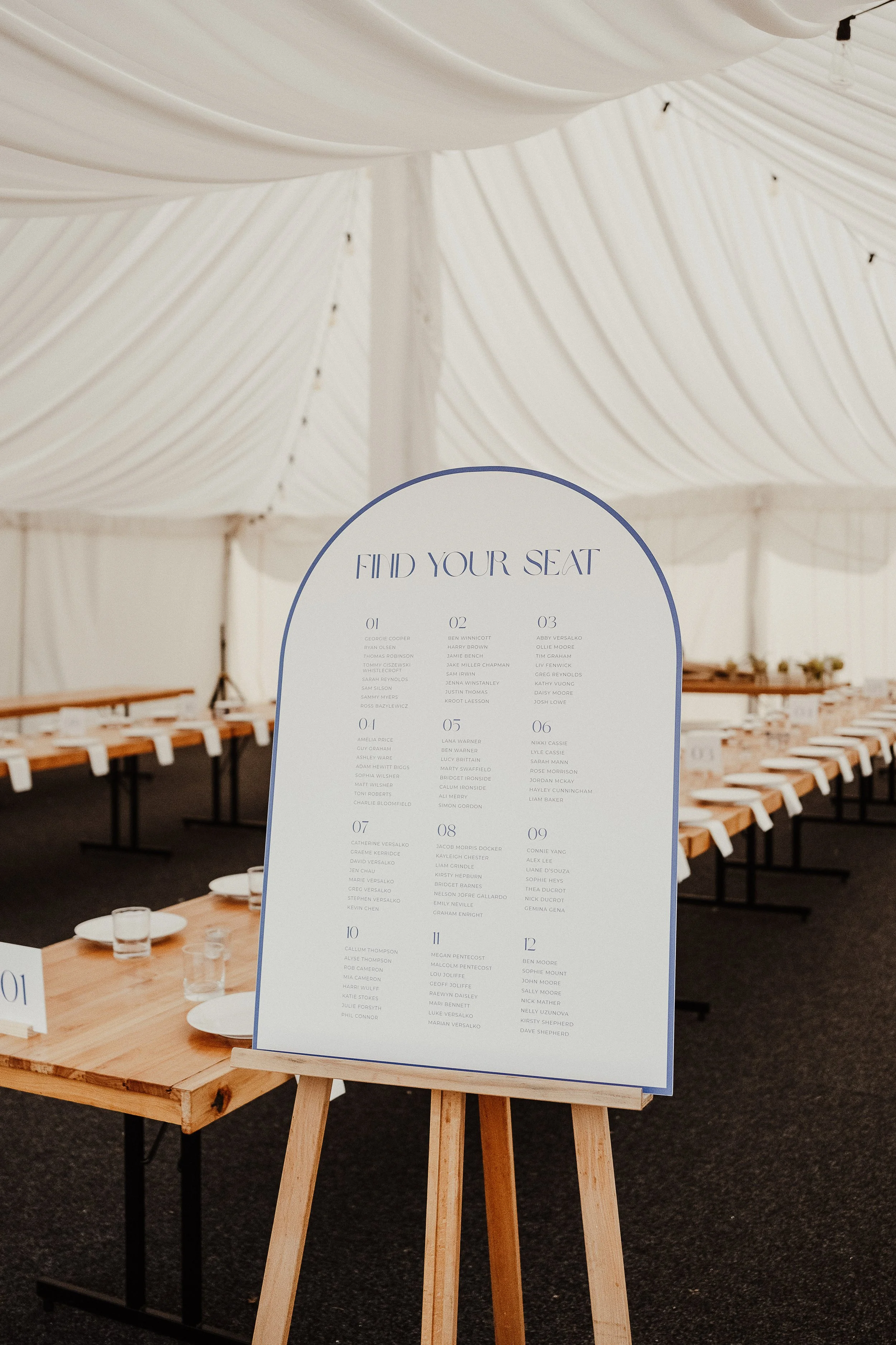 Wedding venue with a table and a seating chart on an easel. The seating chart is titled 'FIND YOUR SEAT' and lists names numbered from 1 to 12. The venue has a white tent with draped fabric on the ceiling and wooden benches around the tables.
