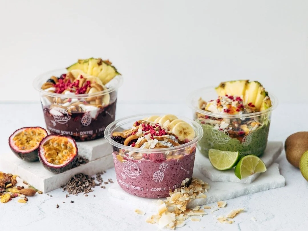 Three colorful smoothie bowls topped with fruit, nuts, and toppings on white marble slabs with passion fruits, lime wedges, and kiwis around them.