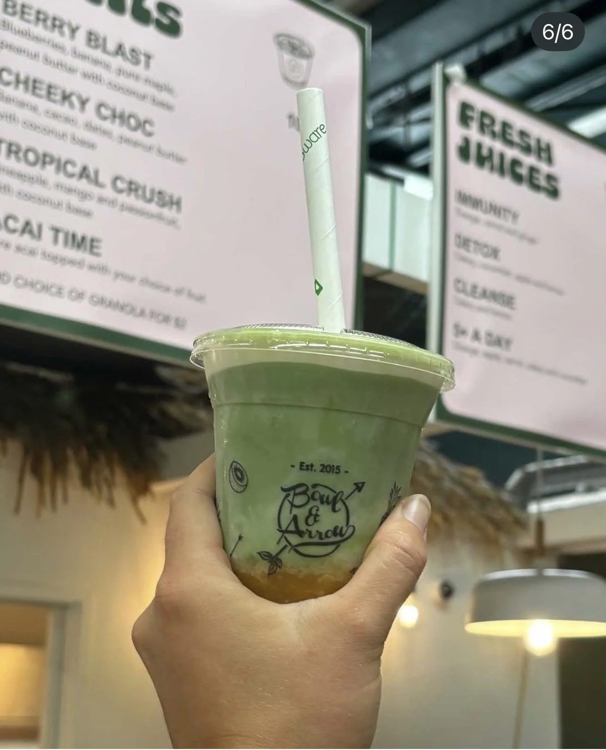 A hand holding a green matcha drink in a clear plastic cup with a sticker and a white straw, in front of menu boards at a juice shop.