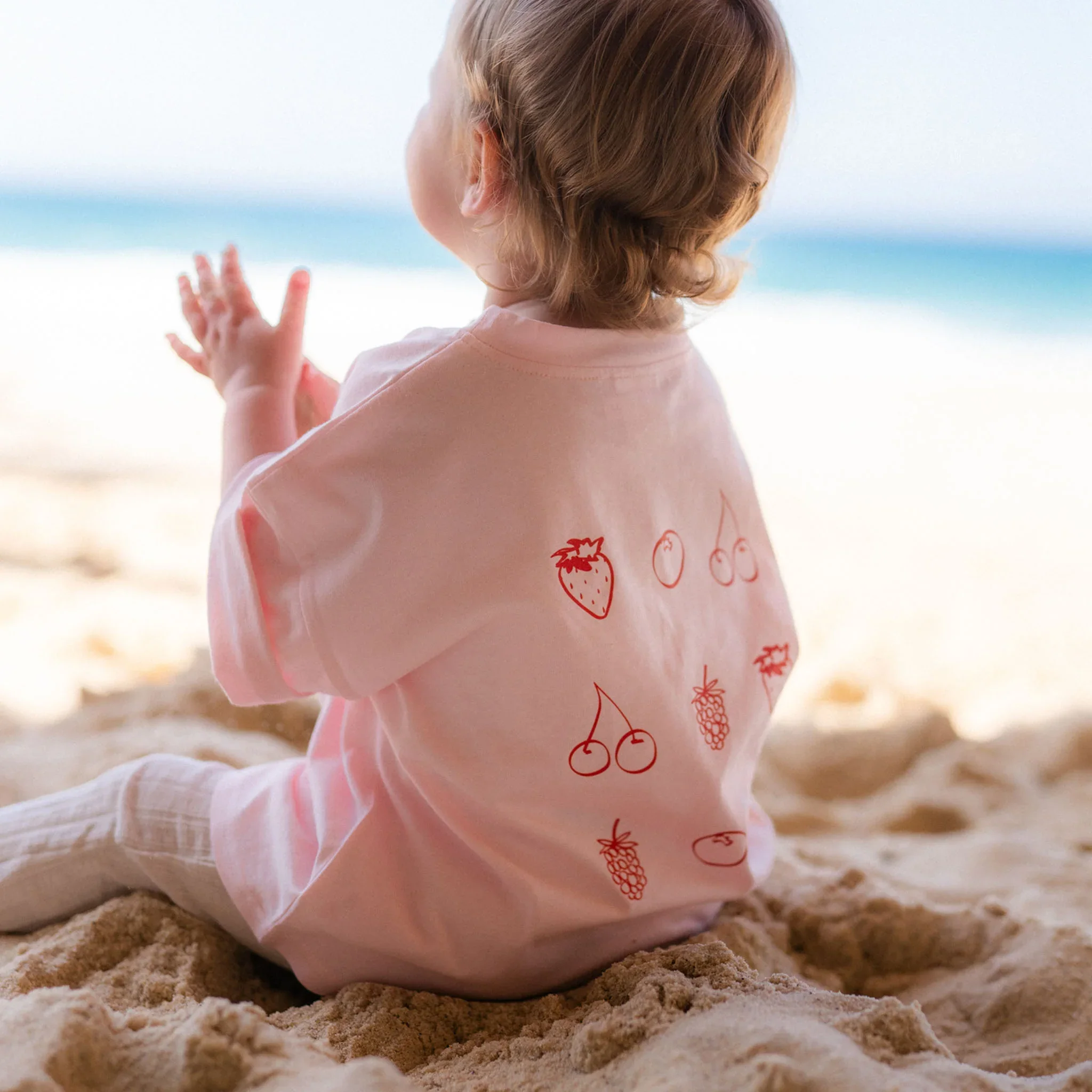 A young child with curly hair wearing a light pink shirt with strawberries and cherries, sitting on sand at the beach, facing away, with ocean and sky in the background.