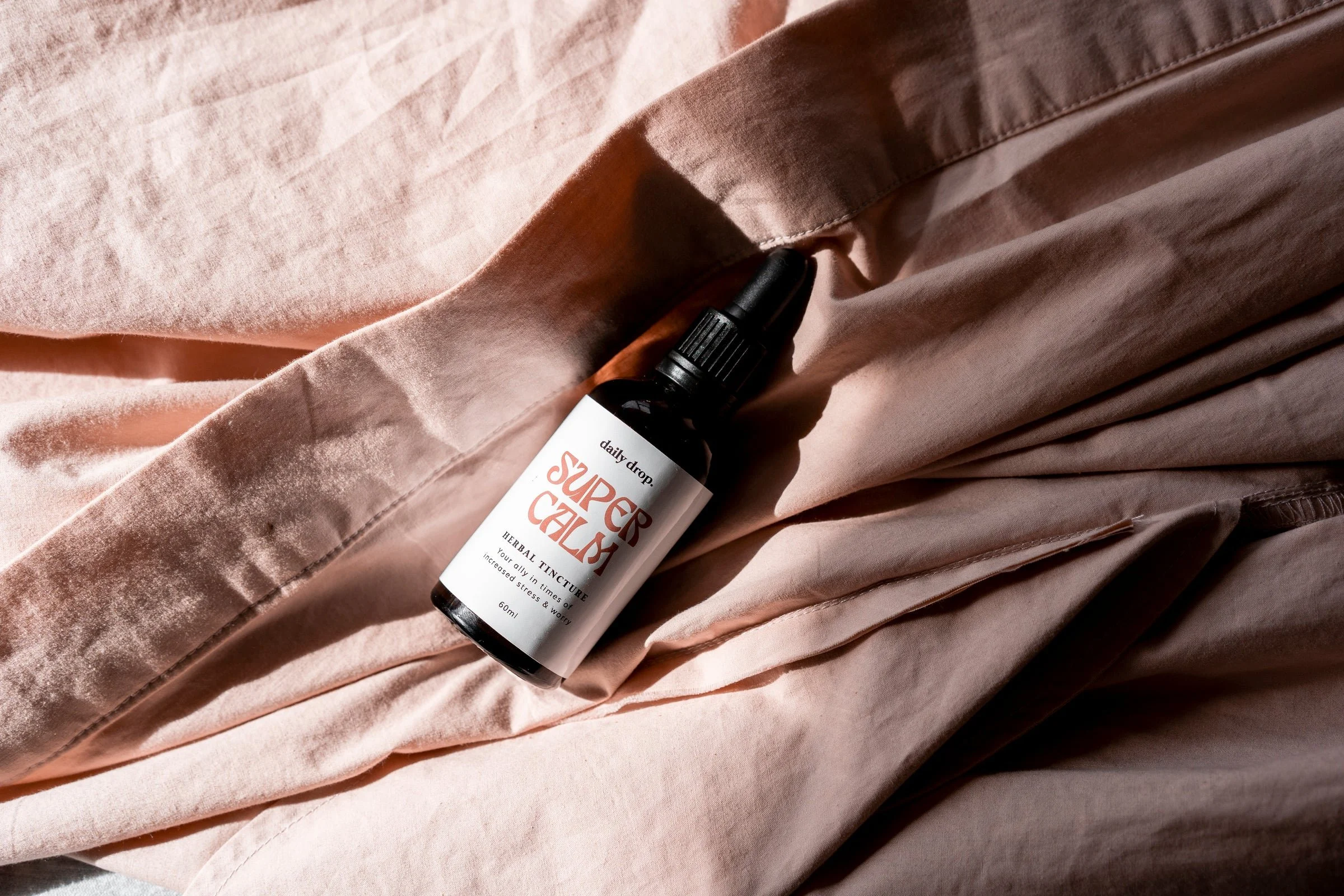 A small dark bottle with a spray nozzle lying on pinkish fabric. The label on the bottle reads 'Super Sale' and mentions herbal tincture with benefits for stress and worry.