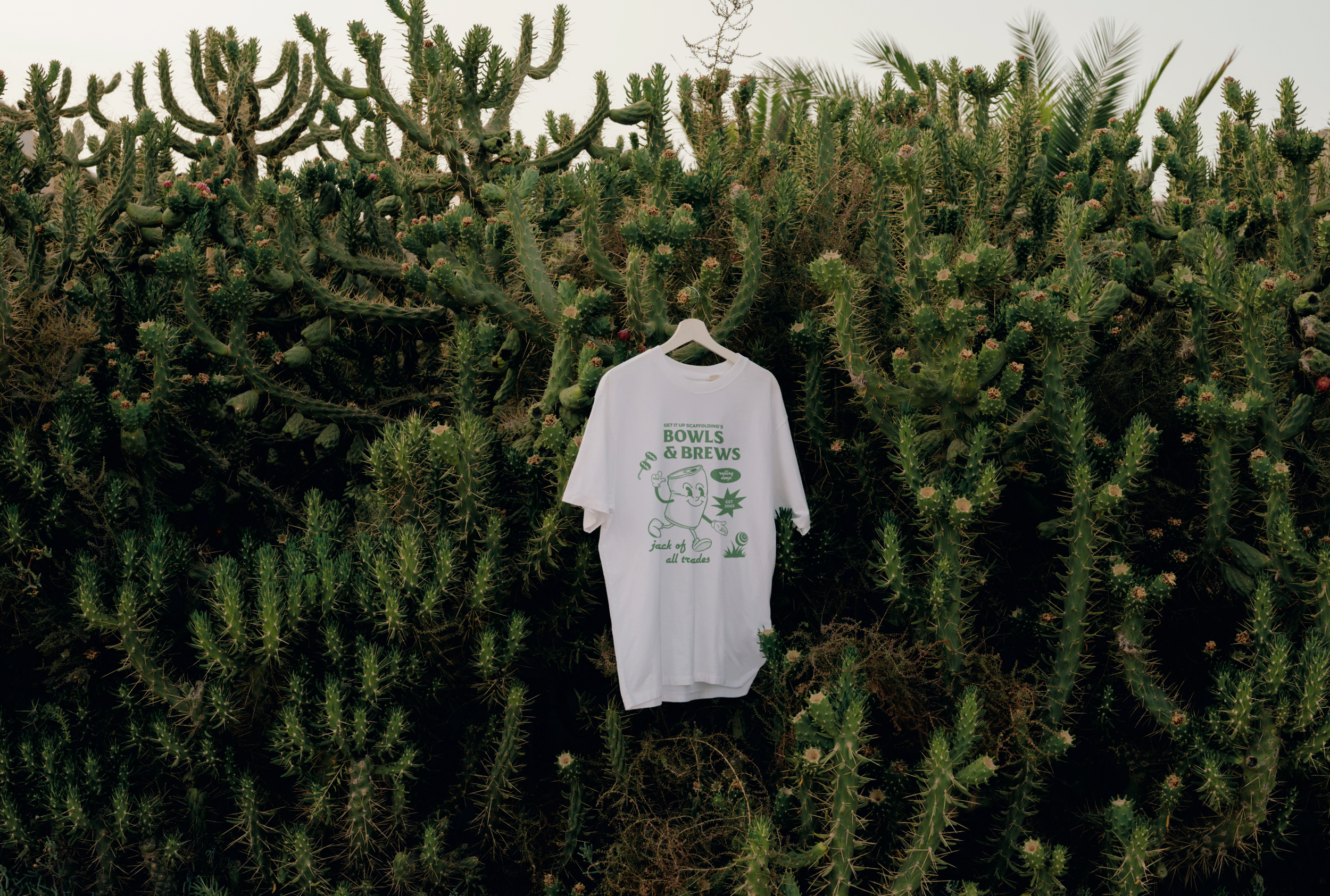 A white T-shirt with green text and a cartoon graphic hangs on a white hanger in front of a dense thicket of green cacti. The T-shirt features text that reads 'Bowls & Brews' and a cartoon character holding a bowl, with additional smaller text underneath.