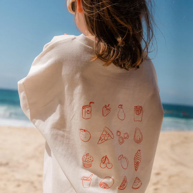 Back of a person wearing a white T-shirt with red illustrated food and drink icons, standing on a beach with ocean and sky in the background.