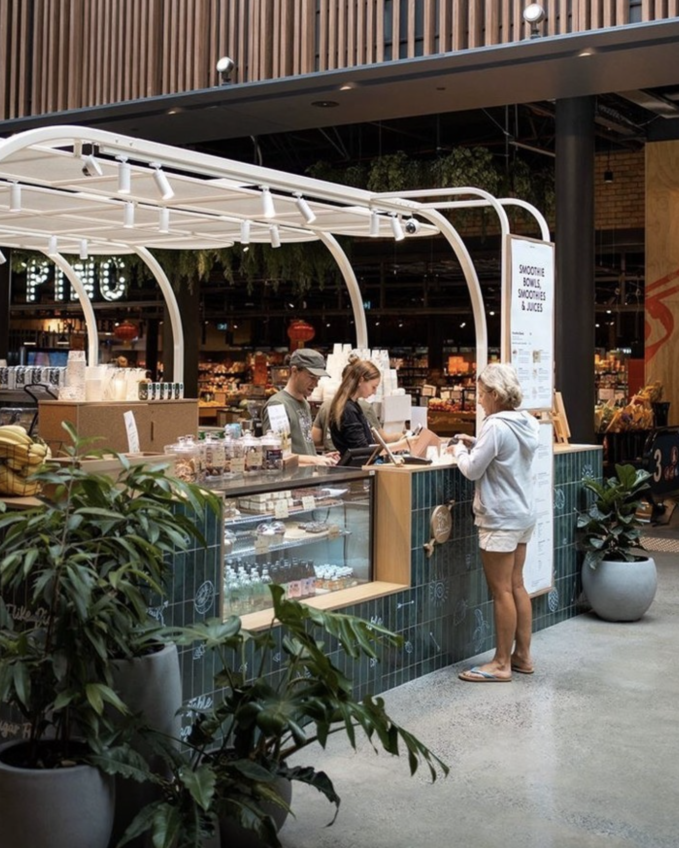 A woman placing an order at a smoothie and juice kiosk inside a modern grocery store, with two staff members behind the counter preparing drinks, and potted plants in the foreground.
