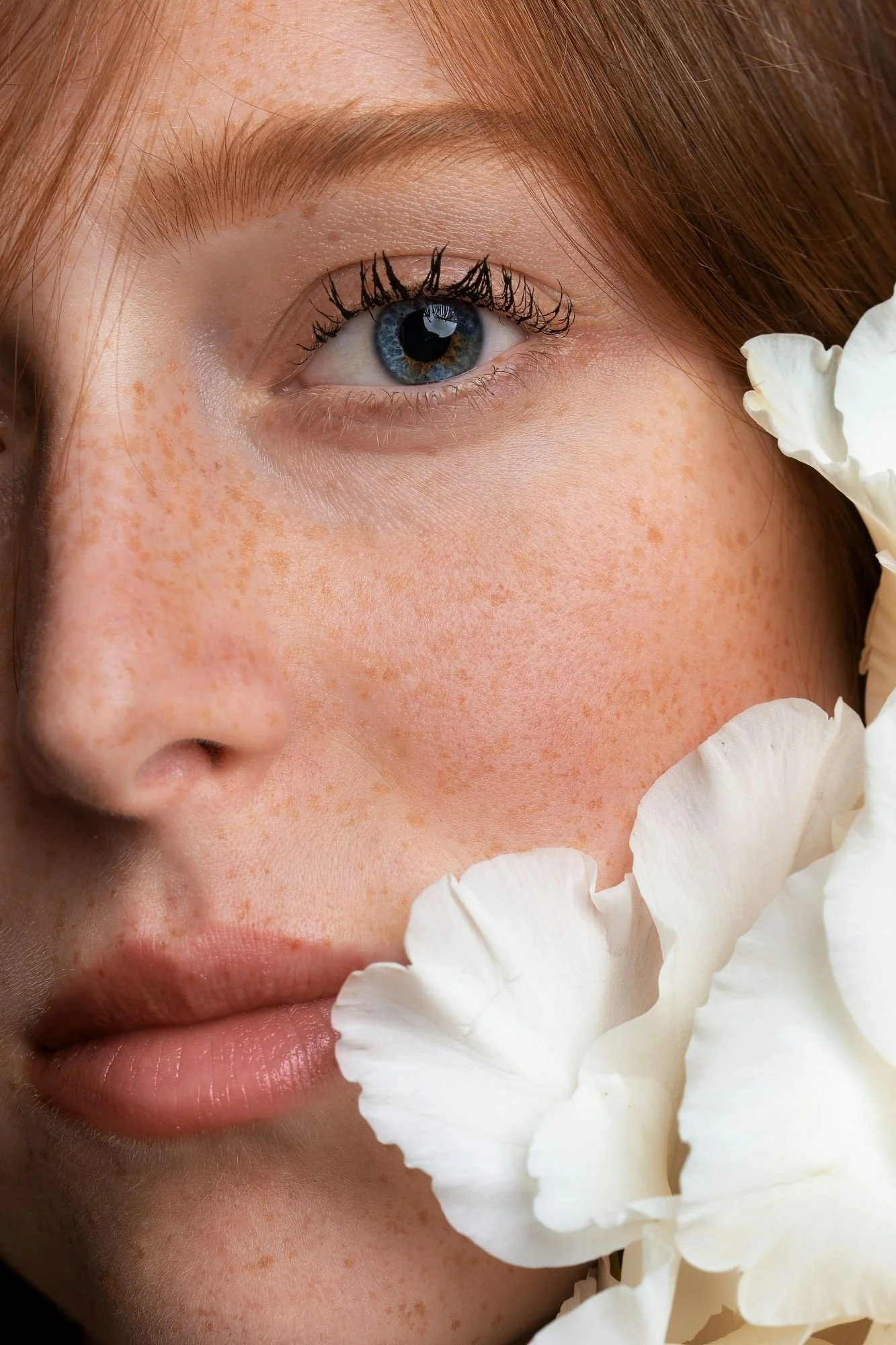 Close-up of a person with red hair, blue eyes, and freckles, surrounded by white flower petals.