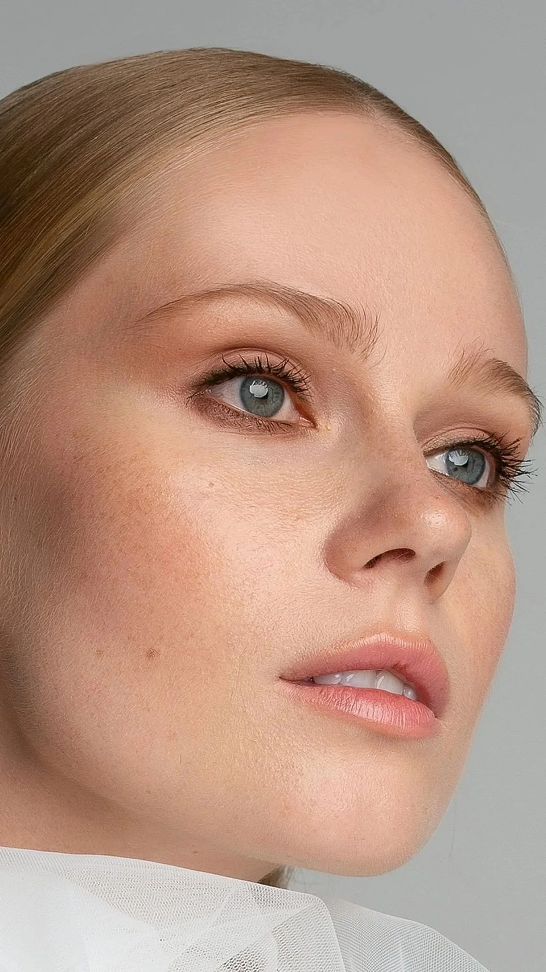 Close-up of a woman's face showing light skin, blue eyes, light eyebrows, and blonde hair pulled back. She has natural makeup with soft pink lips and is wearing a white garment with sheer fabric.