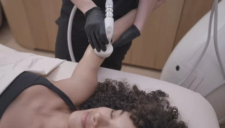 A woman lying on a treatment bed receiving a laser or ultrasound therapy on her arm from a practitioner wearing black gloves in a clinical setting.