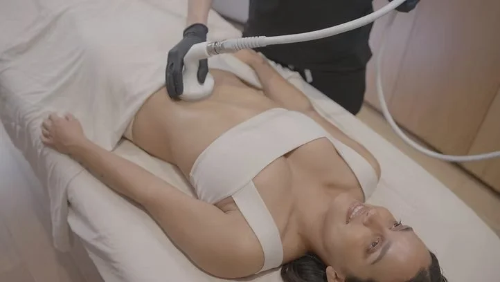 A woman lying on a medical bed receiving a treatment with a handheld device, while a healthcare professional operates the device.