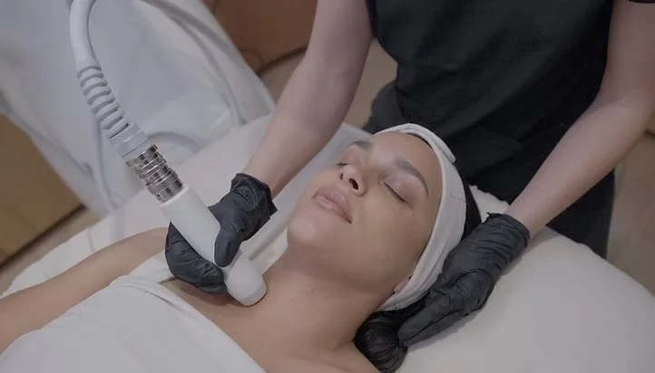 Woman receiving facial treatment with a skincare device, lying on a treatment bed with her eyes closed and head wrapped in a towel, in a professional spa setting.