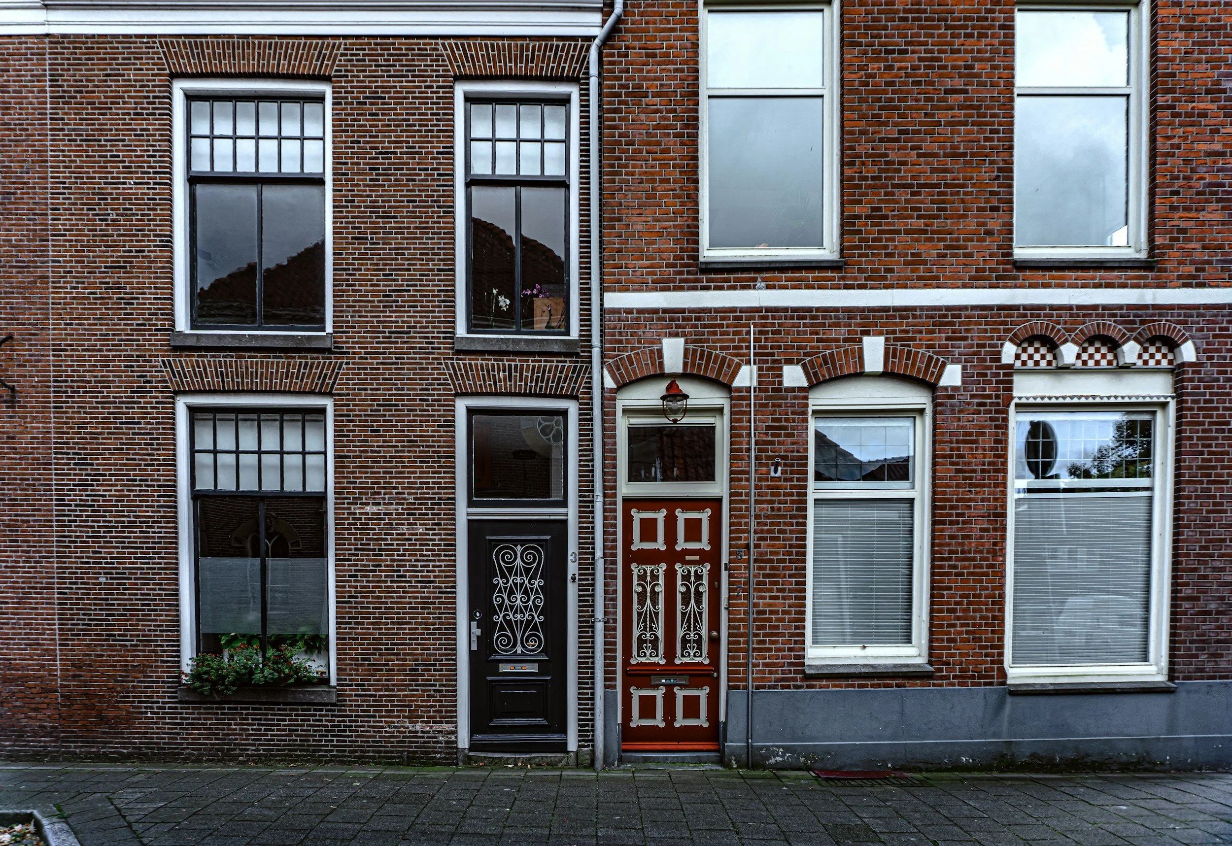 Tweetalige rijtjeshuis met twee verschillende ingangstijlen, een zwarte deur met smeedwerk en een houten deur met rood-wit versteklegging, allemaal met grote ramen en een bakstenen gevel.