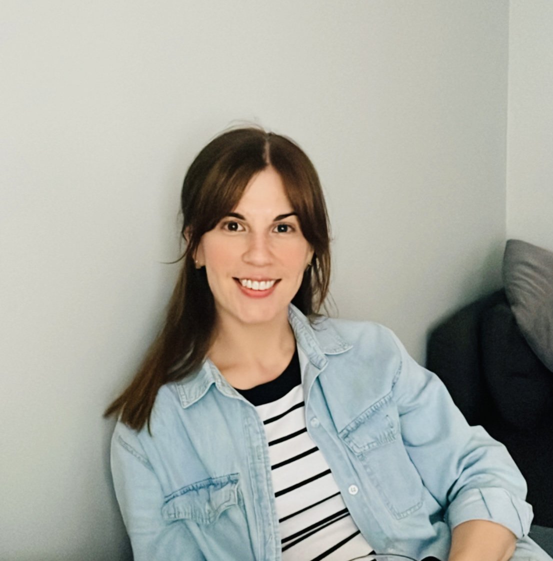 A smiling woman with brown hair, wearing a black and white striped shirt and a light blue denim jacket, sitting on a sofa against a plain wall.