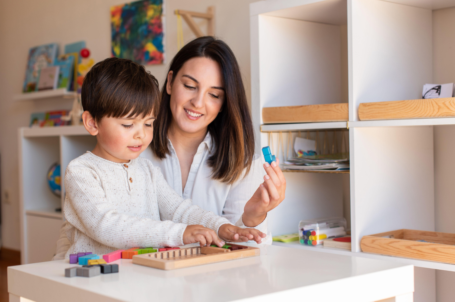 A woman and a young girl sitting at a white table playing with colorful educational toys in a room with shelves filled with books and art.
