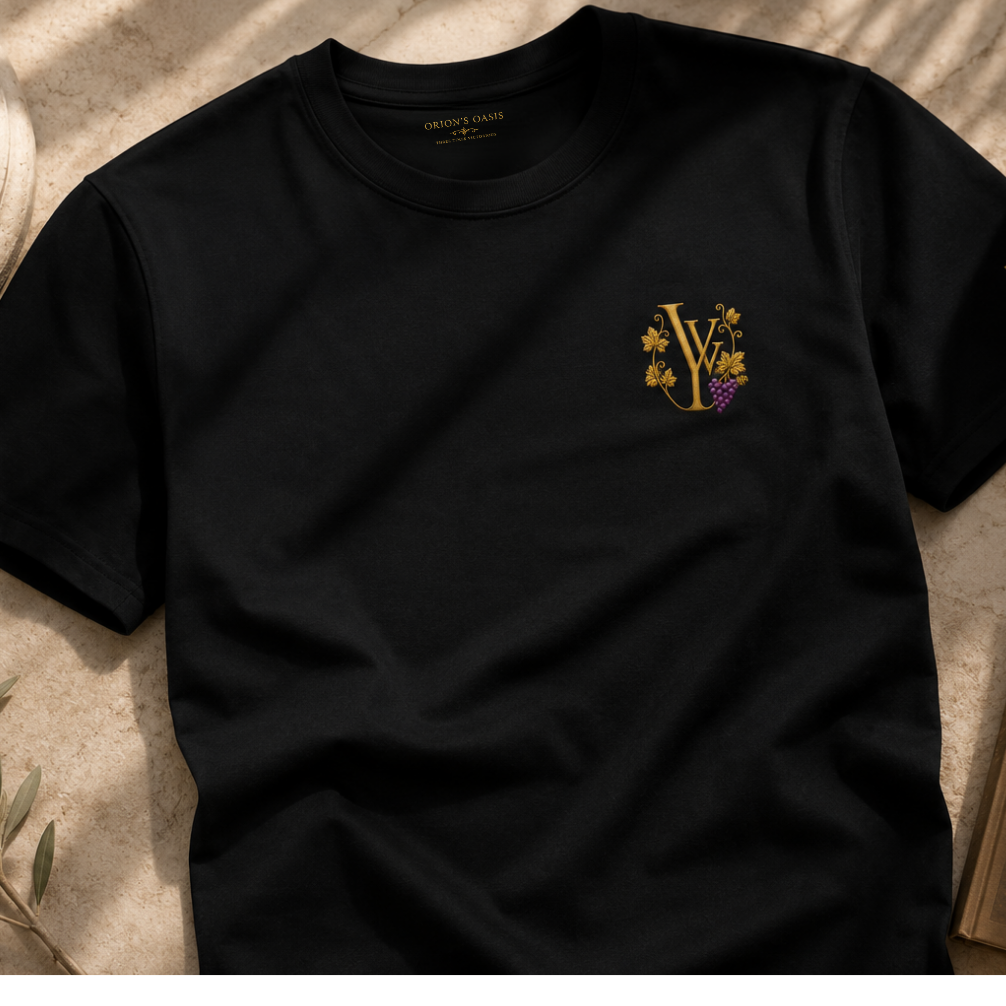 A black crewneck sweatshirt with a gold and purple embroidered logo of grapes and floral design on the chest, laying on a beige marble surface.