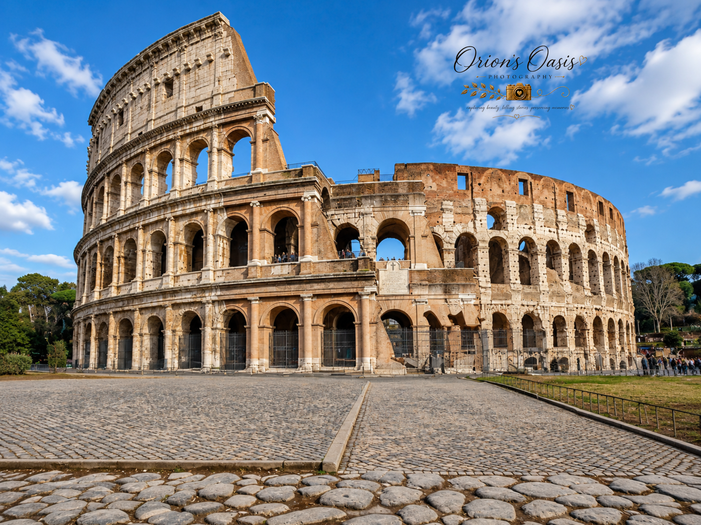 The image shows the ancient Roman Colosseum with a clear blue sky and scattered white clouds overhead. The foreground features a cobblestone street leading to the monument, which is partially surrounded by a fence and visitors can be seen walking around the base.