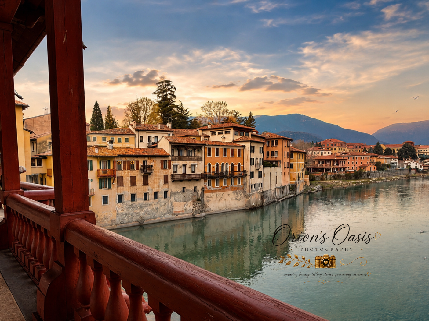 View of colorful buildings along a river at sunset, with mountains in the background, seen from a balcony with a red railing.