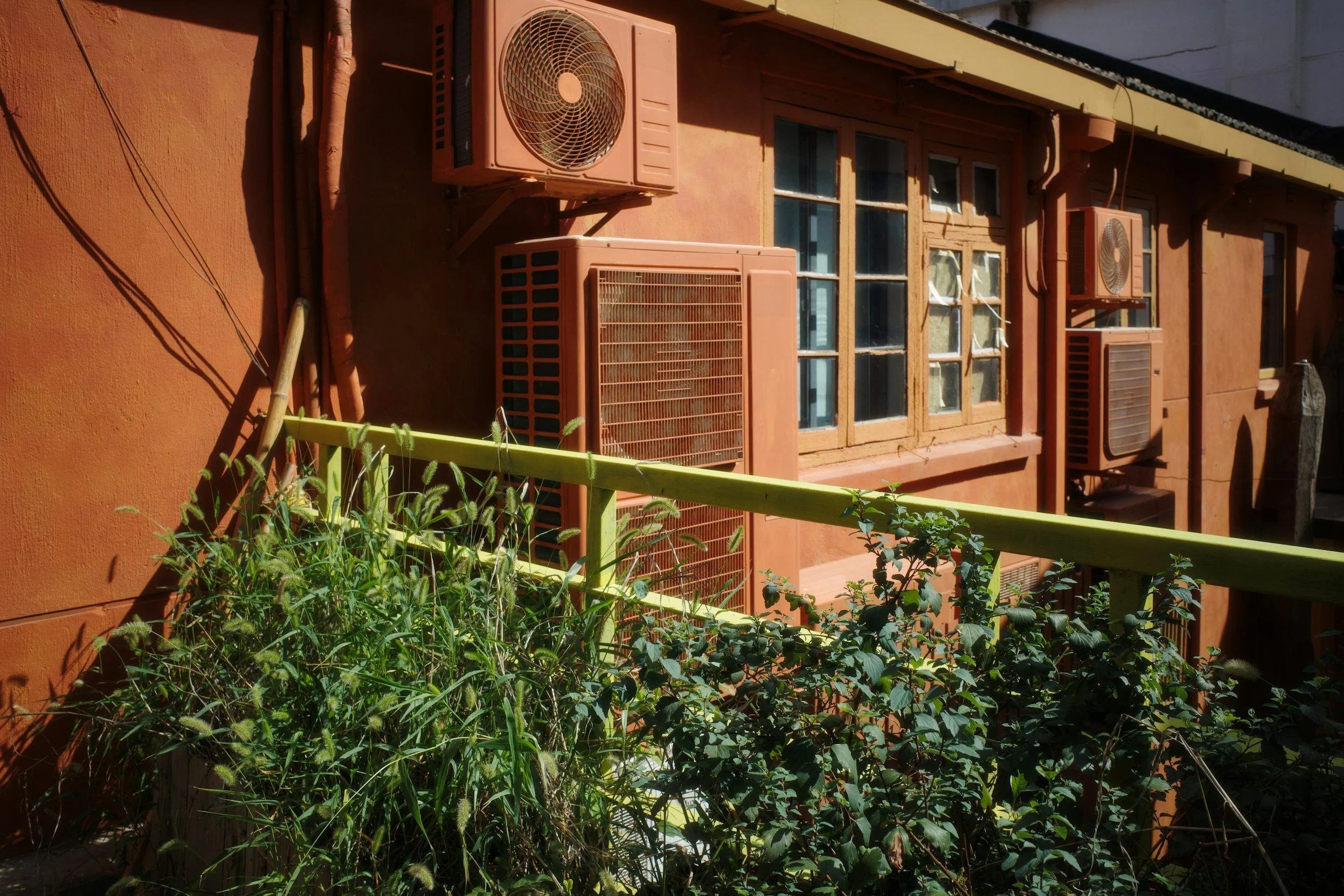 Exterior of a building with multiple air conditioning units mounted on the wall, a window with small panes, and green plants in the foreground.