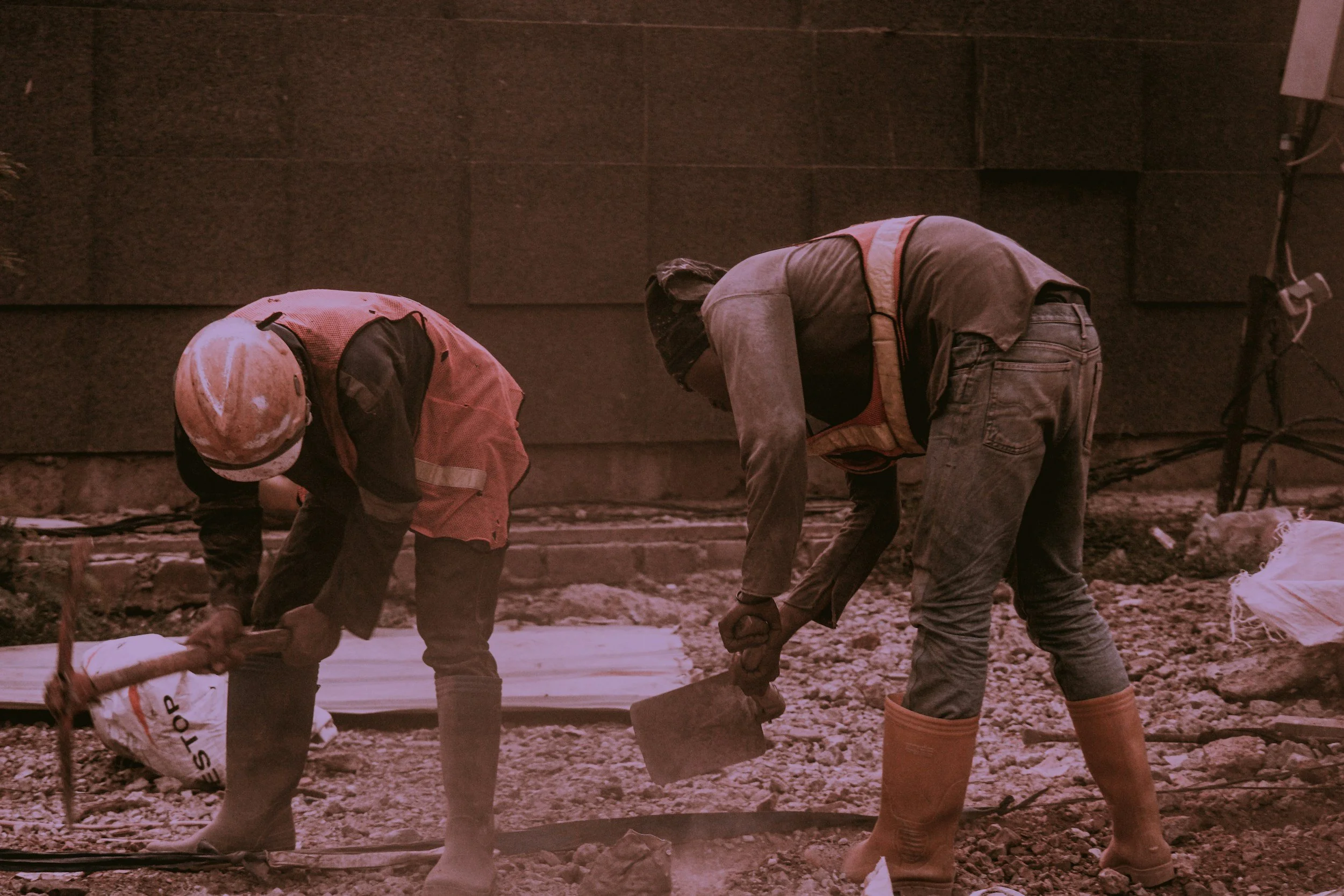 Two construction workers bending over working on ground, one holding a bag labeled 'STOP' and the other with a saw, at building site with dark brick wall background.