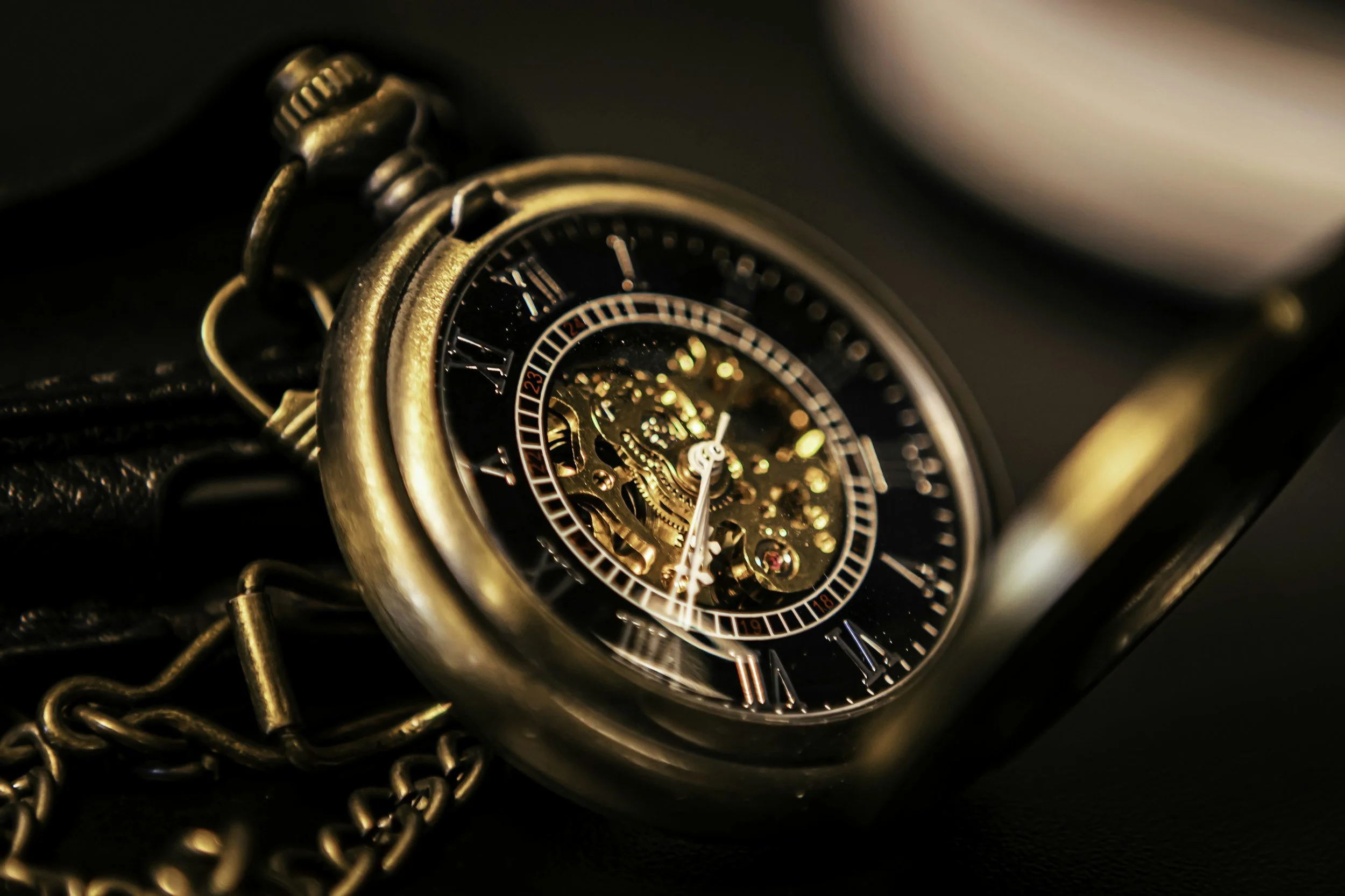 A close-up image of a vintage-style watch with a chain, showing intricate gears and Roman numeral markings on the dial, with a dark background.