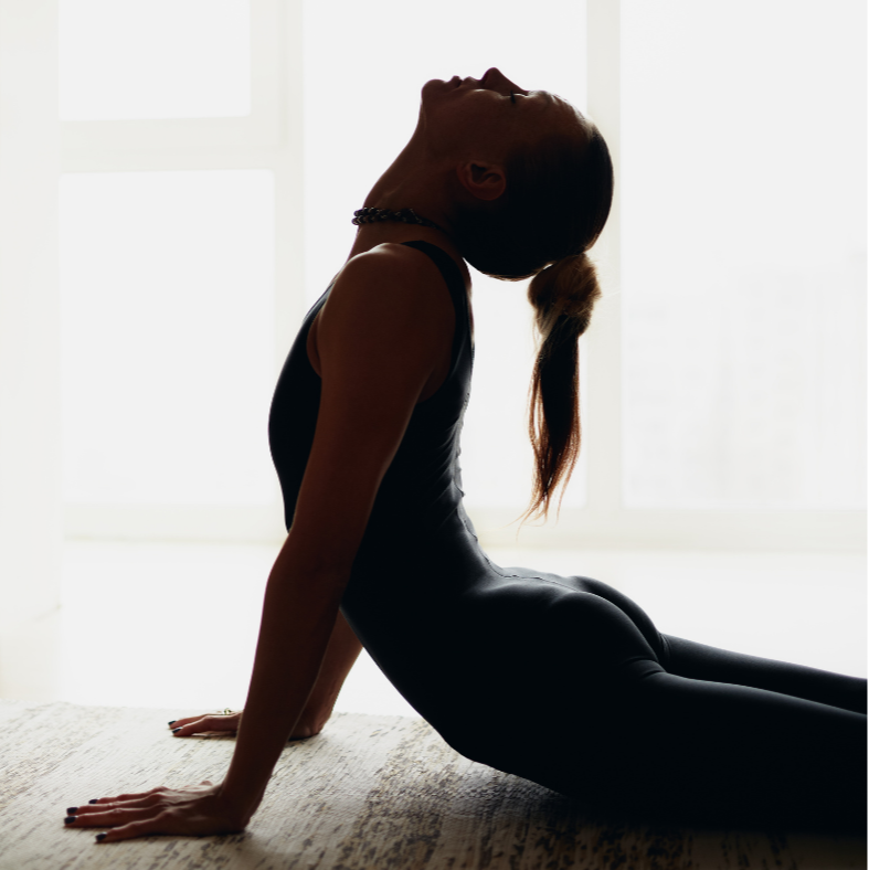 A woman wearing black workout clothes and a necklace, performing a yoga pose on a textured surface with backlit windows in the background.