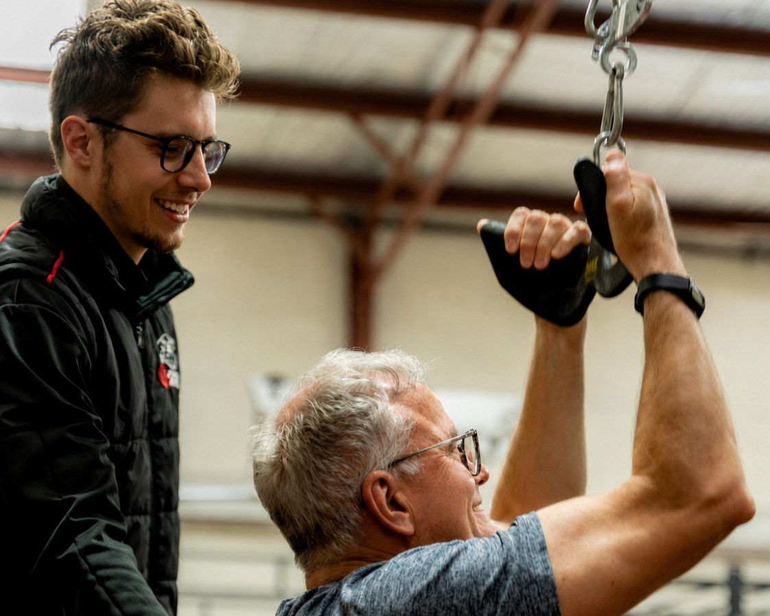 A young trainer or coach assisting an older man with a weightlifting exercise in a gym.