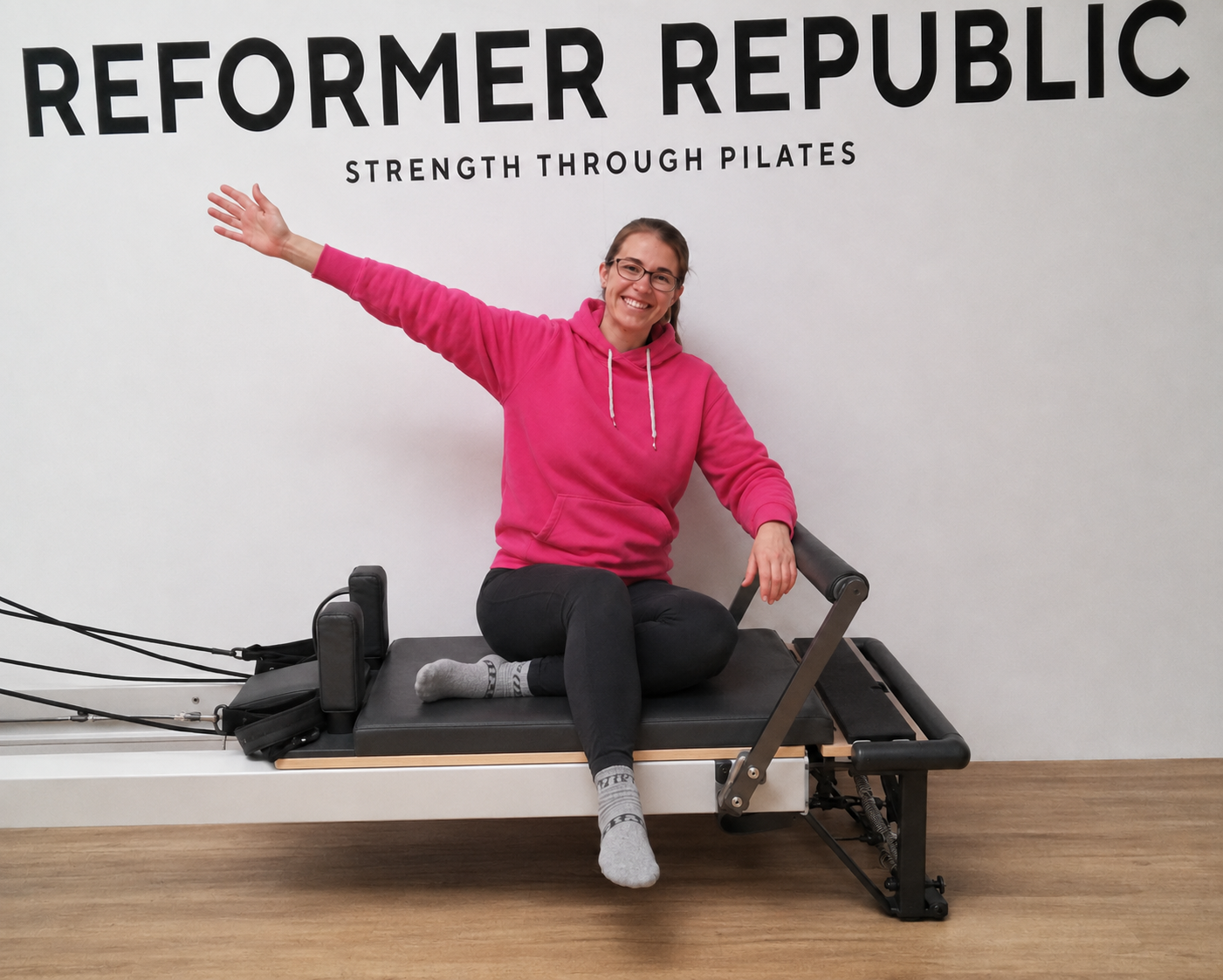 A woman in a pink hoodie sits on a reformer Pilates machine with one arm extended to the side and a smile. The wall behind her has the words 'Reformer Republic' and 'Strength Through Pilates' in bold black letters.