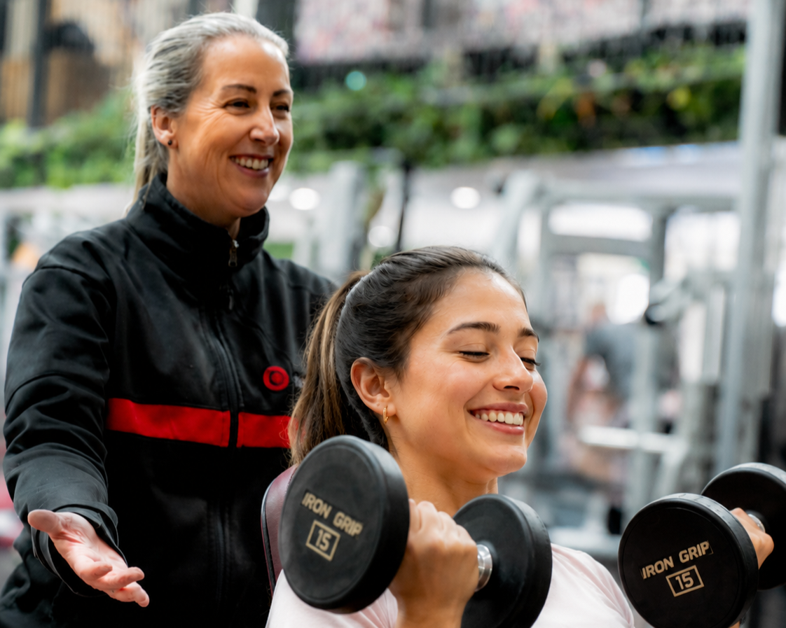 A woman is working out with dumbbells while a trainer encourages her at the gym.