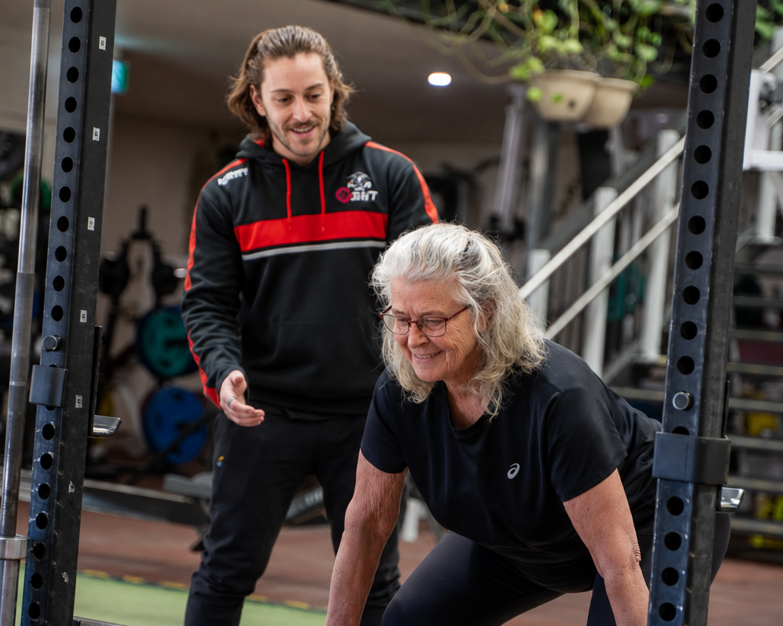 An elderly woman doing a push-up with a man mentoring her in a gym.