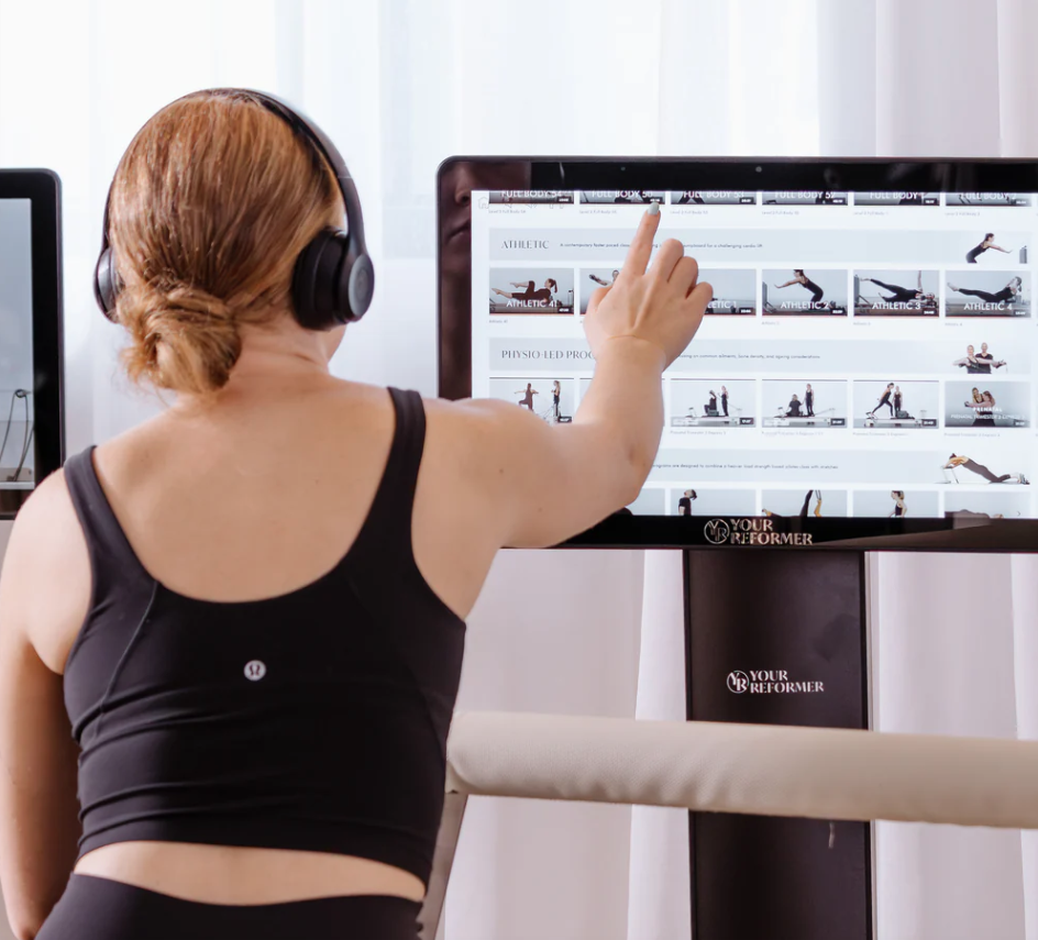 Person with red hair wearing headphones and a black athletic top pointing at exercise images on a large screen