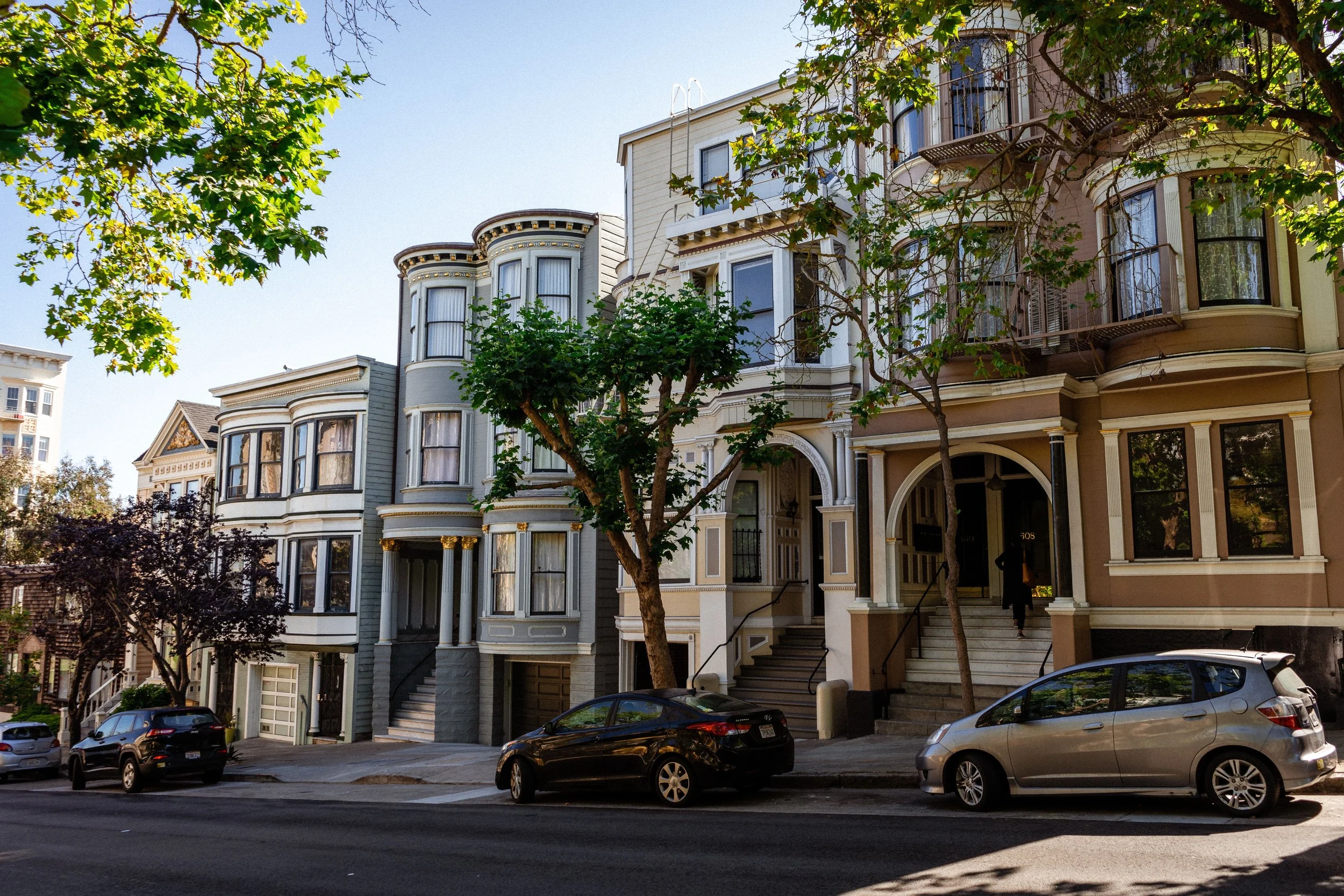 Historic Victorian row houses with ornate bay windows and entryways lining a tree-lined city street with parked cars