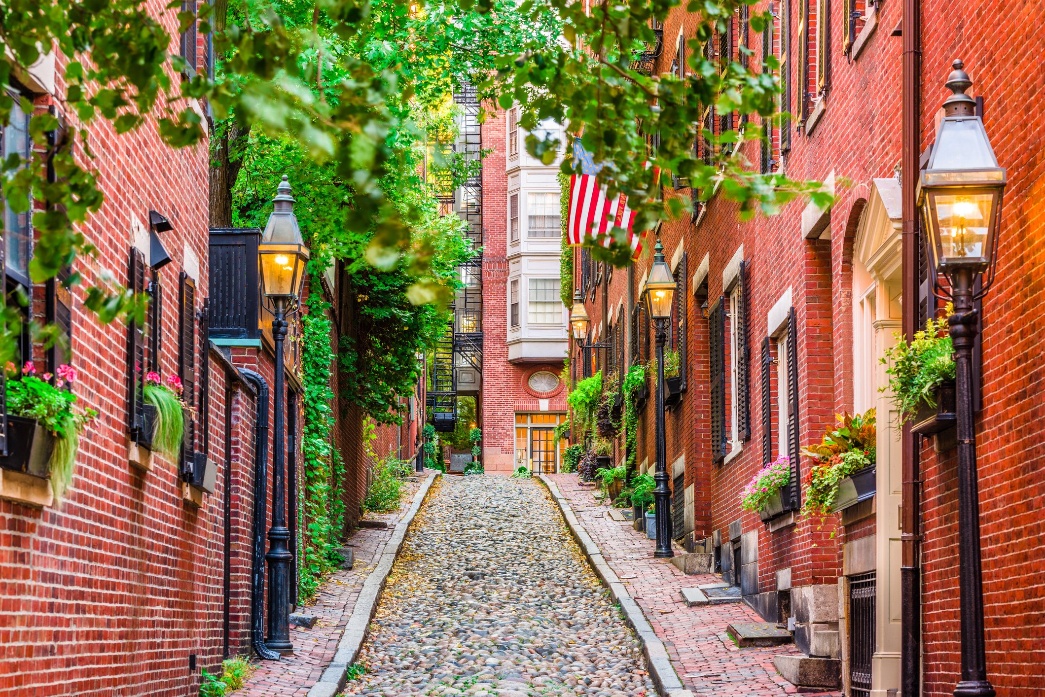 Historic Boston cobblestone alley lined with red brick homes, greenery, lanterns, and an American flag
