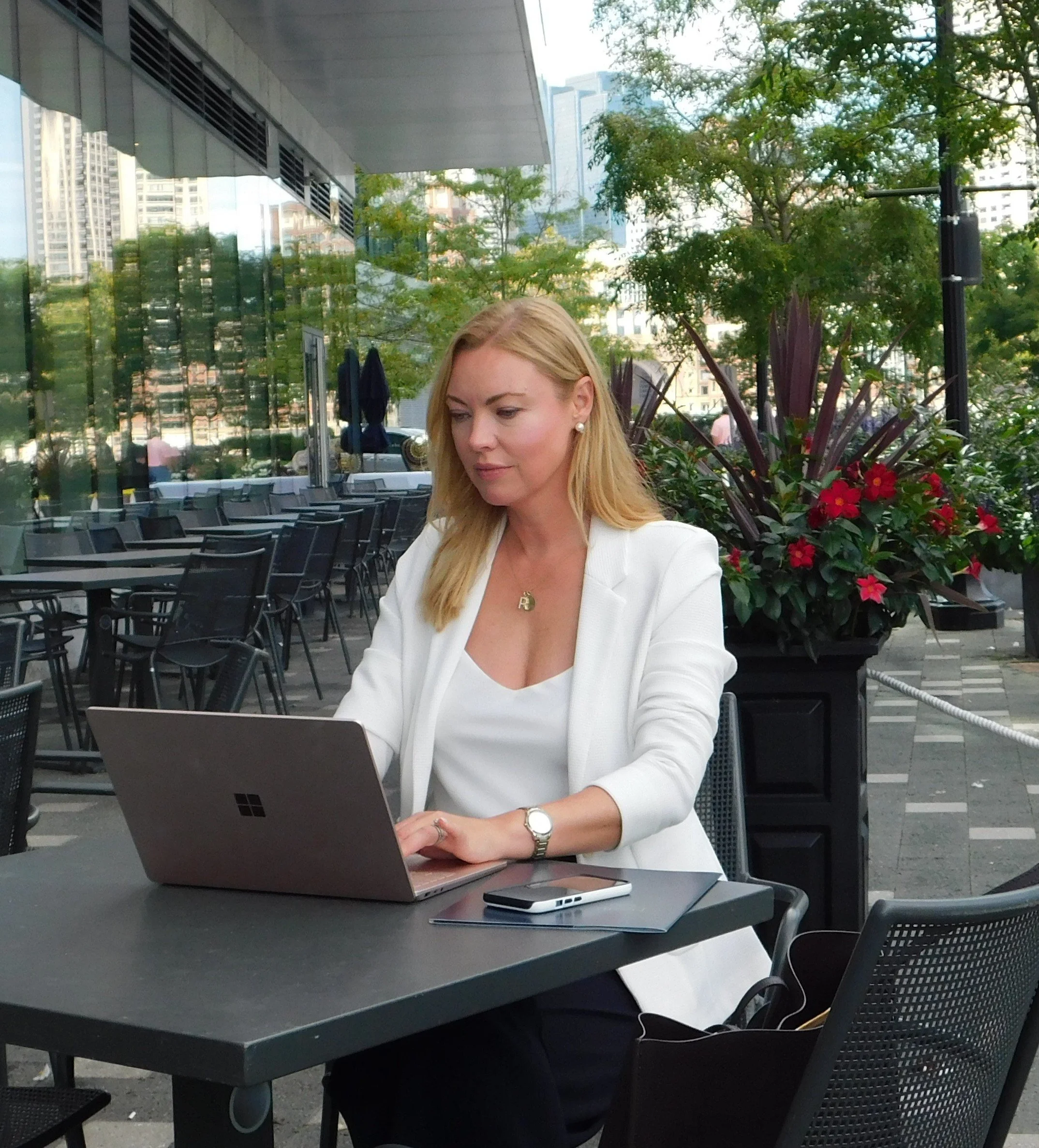 Woman working on a laptop at an outdoor cafe with city buildings and plants in the background