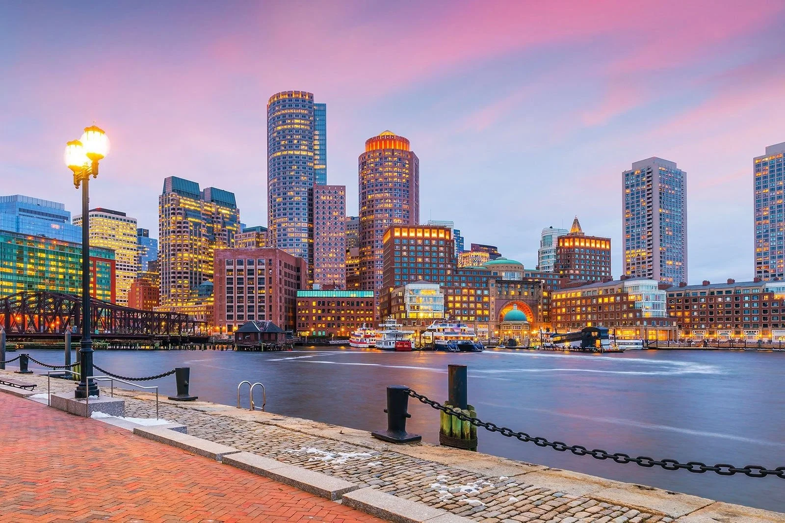 Boston skyline at dusk viewed from the waterfront, with illuminated skyscrapers, boats, and harbor promenade