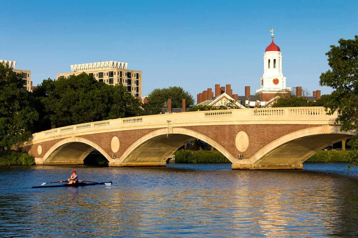 Rower on the Charles River beneath a historic arched bridge with Boston skyline and church steeple