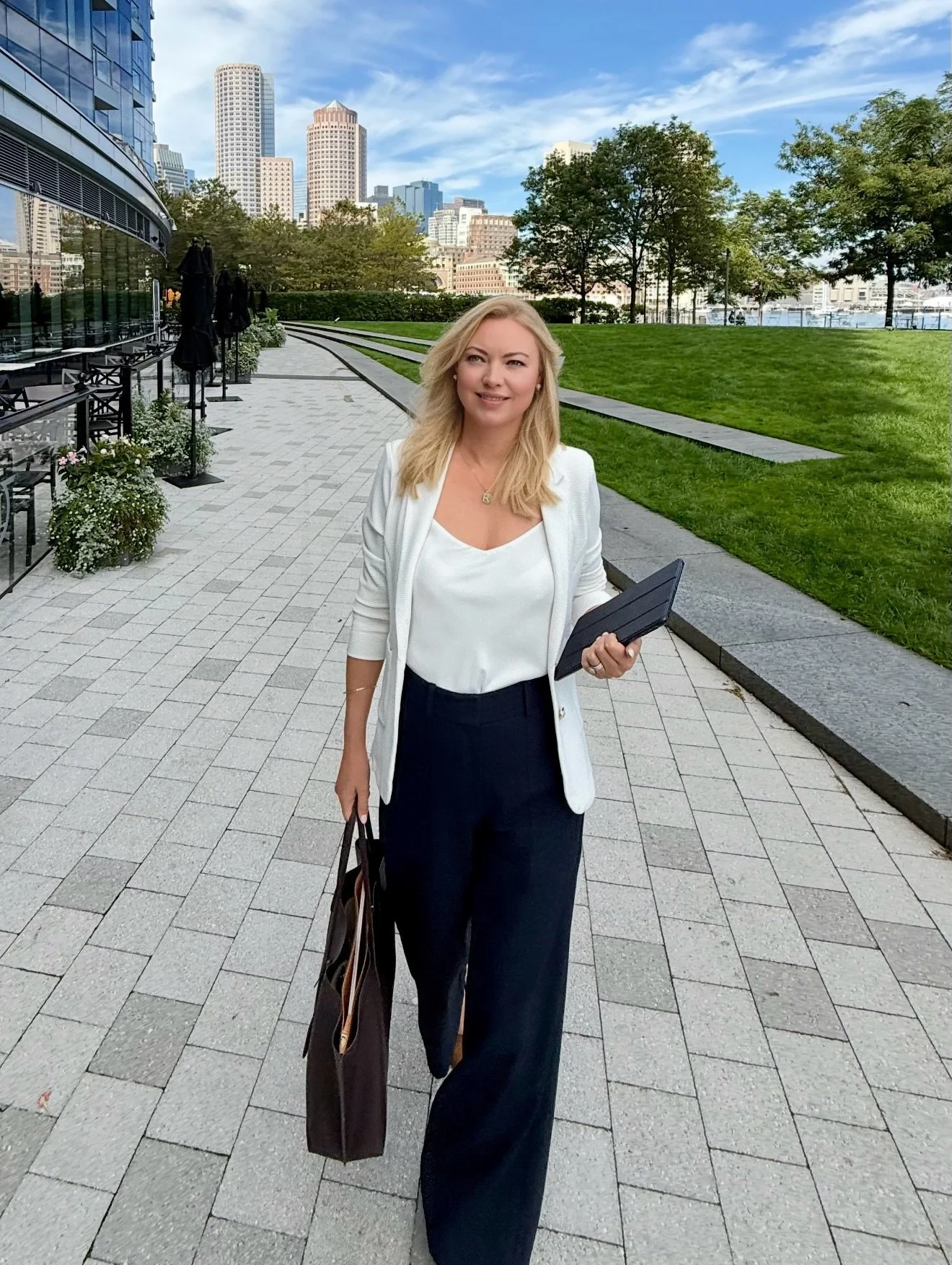 Blonde businesswoman holding a tablet and tote bag on a waterfront promenade with a city skyline behind her