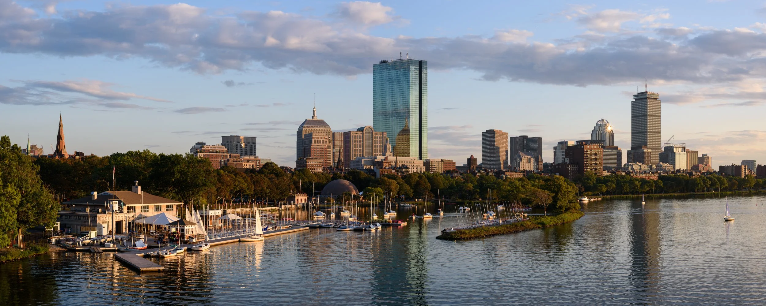 Boston skyline at sunset viewed across the Charles River, with sailboats in the foreground and Boston Common landmarks including the Prudential Tower and 200 Clarendon Street.
