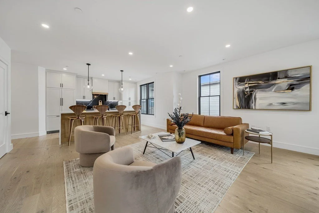 Modern open-concept living room and kitchen with brown sofa, upholstered chairs, island bar seating, hardwood floors, area rug, and abstract wall art